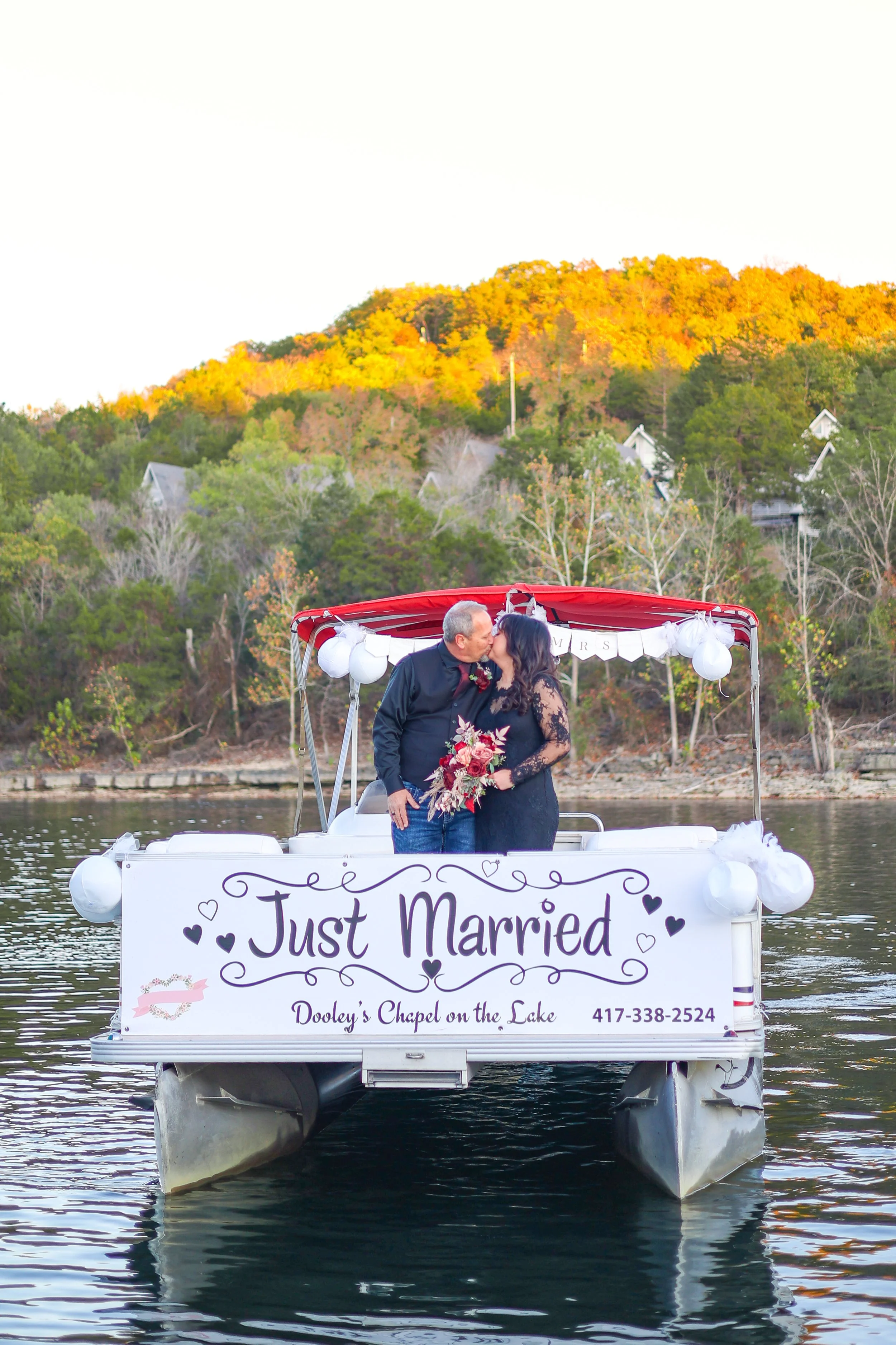 A newly married couple kissing on a boat decorated with a 'Just Married' sign, floating on a lake with a wooded hillside in the background at sunset.