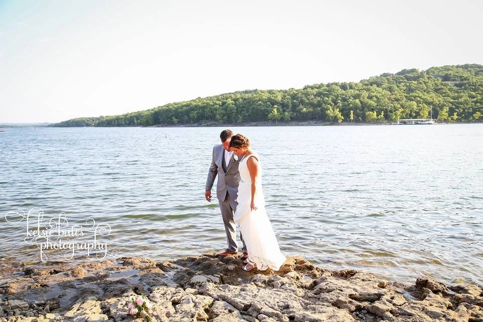 A bride and groom standing on rocky shoreline by a lake with a green hillside in the background, during sunset