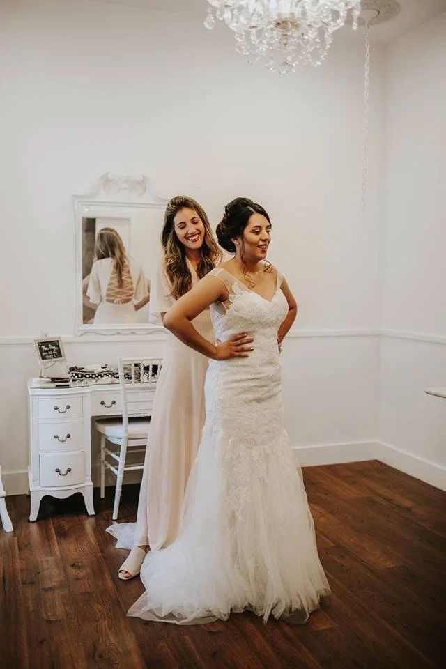 Two women, one in a wedding dress and the other in a light-colored dress, smiling in a room with white walls and wooden floors, near a mirror and a small white desk.