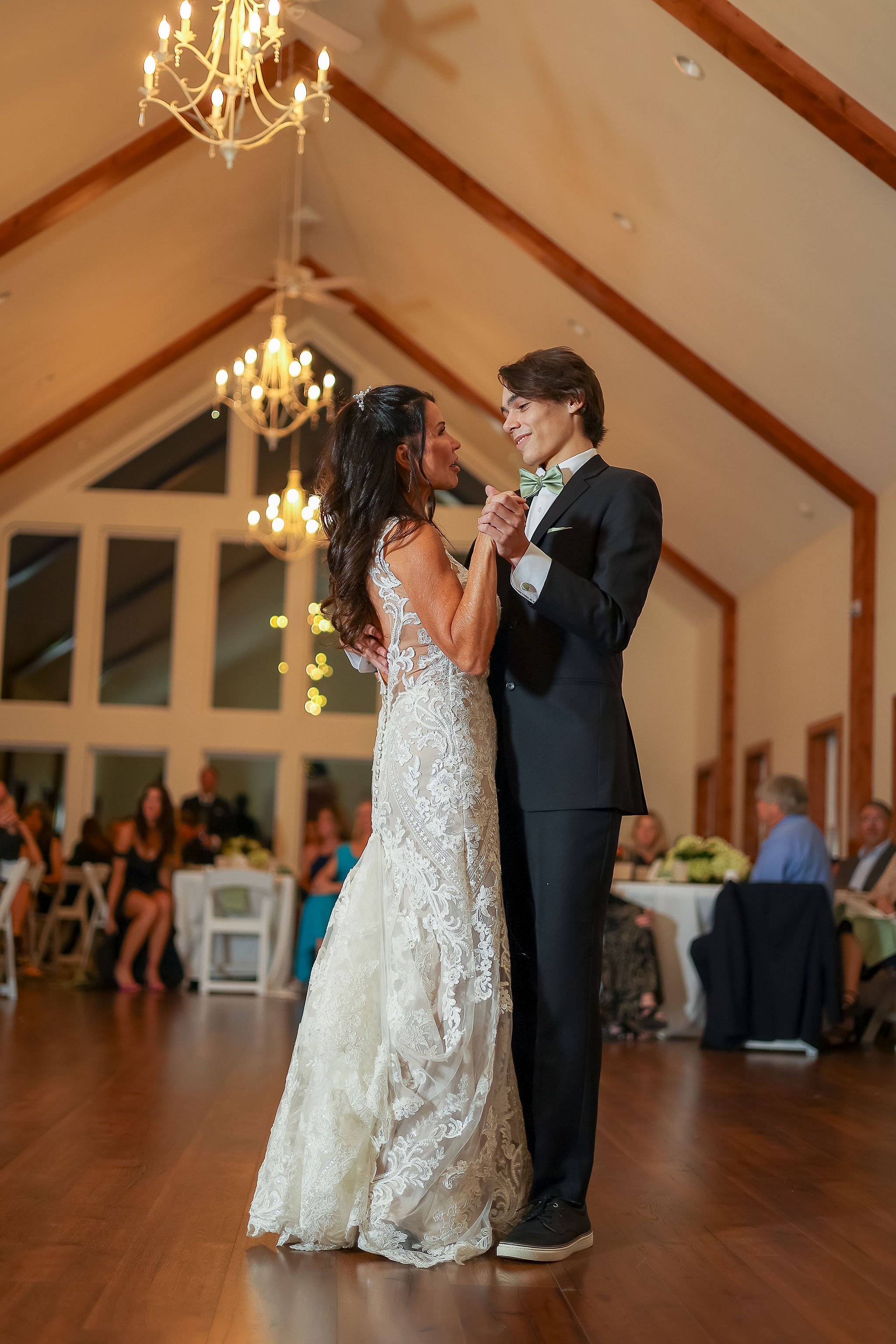 A bride and groom are dancing together during their wedding reception in a warmly lit room with chandeliers and large windows.