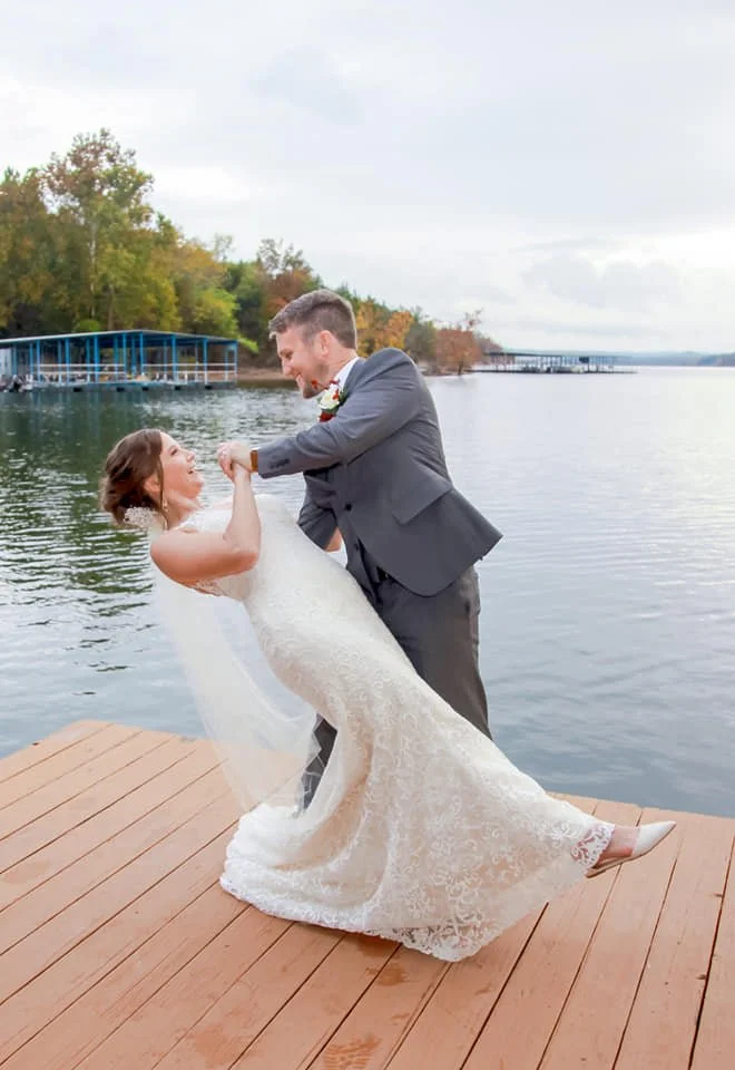 A newlywed couple dancing on a wooden dock by a river, with trees and a pier in the background.
