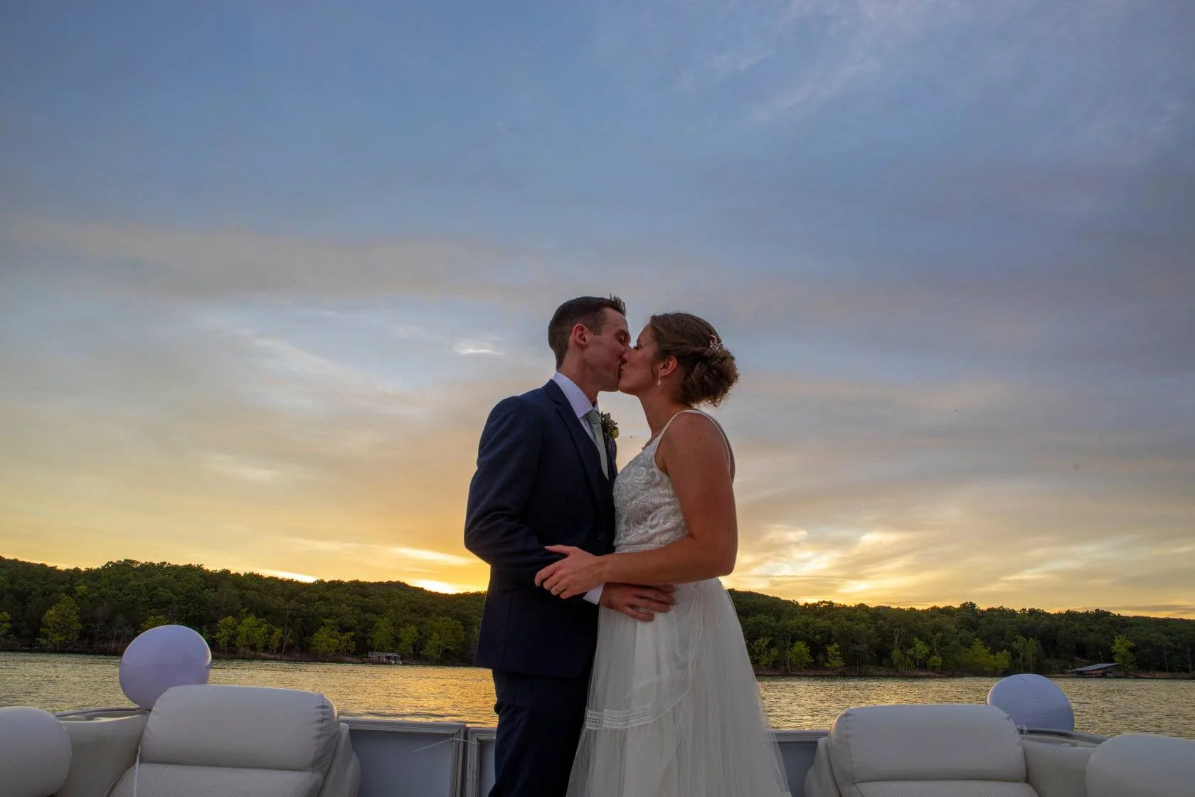 A couple in wedding attire kissing on a boat at sunset with a lake and green trees in the background.