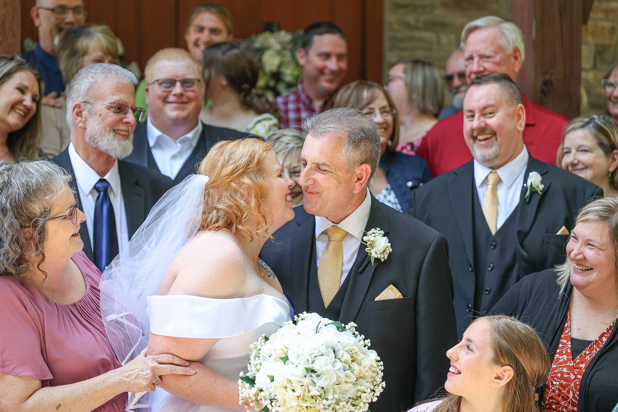 A bride and groom appear to be exchanging a kiss during a wedding ceremony, surrounded by smiling family and friends in a warmly decorated indoor or outdoor venue.
