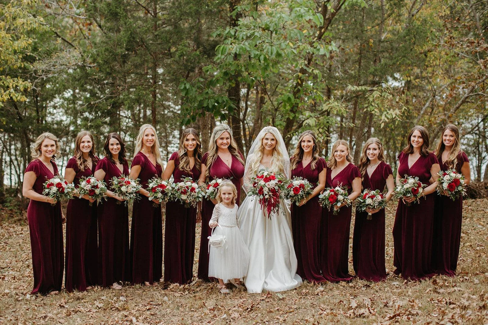 Group of women in matching maroon dresses, holding bouquets of flowers, standing outdoors in a wooded area with fallen leaves, with a bride in a white gown and veil holding a large floral bouquet, and a young girl in a white dress in front of her.