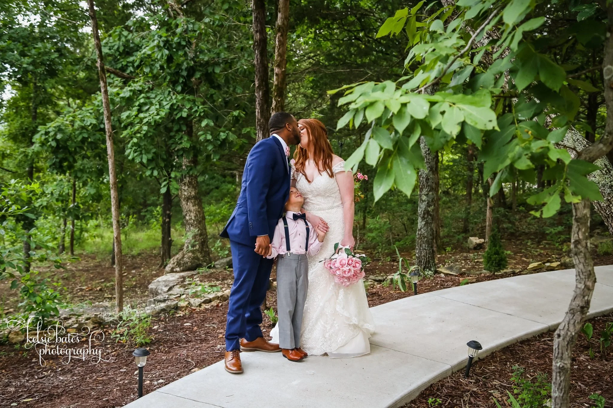 A couple kissing at their wedding in a forest, with a young boy holding their hands, on a concrete pathway surrounded by trees and greenery.
