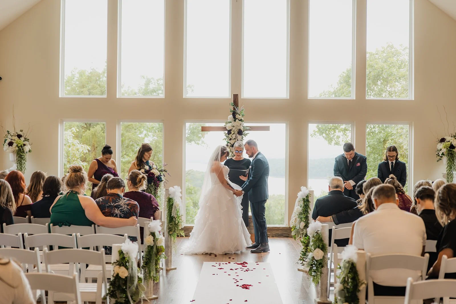 A wedding ceremony taking place in a bright room with large windows. The bride and groom stand at the altar, holding hands and facing each other, with a person officiating behind them. Guests sit in white chairs decorated with flowers, and rose petal