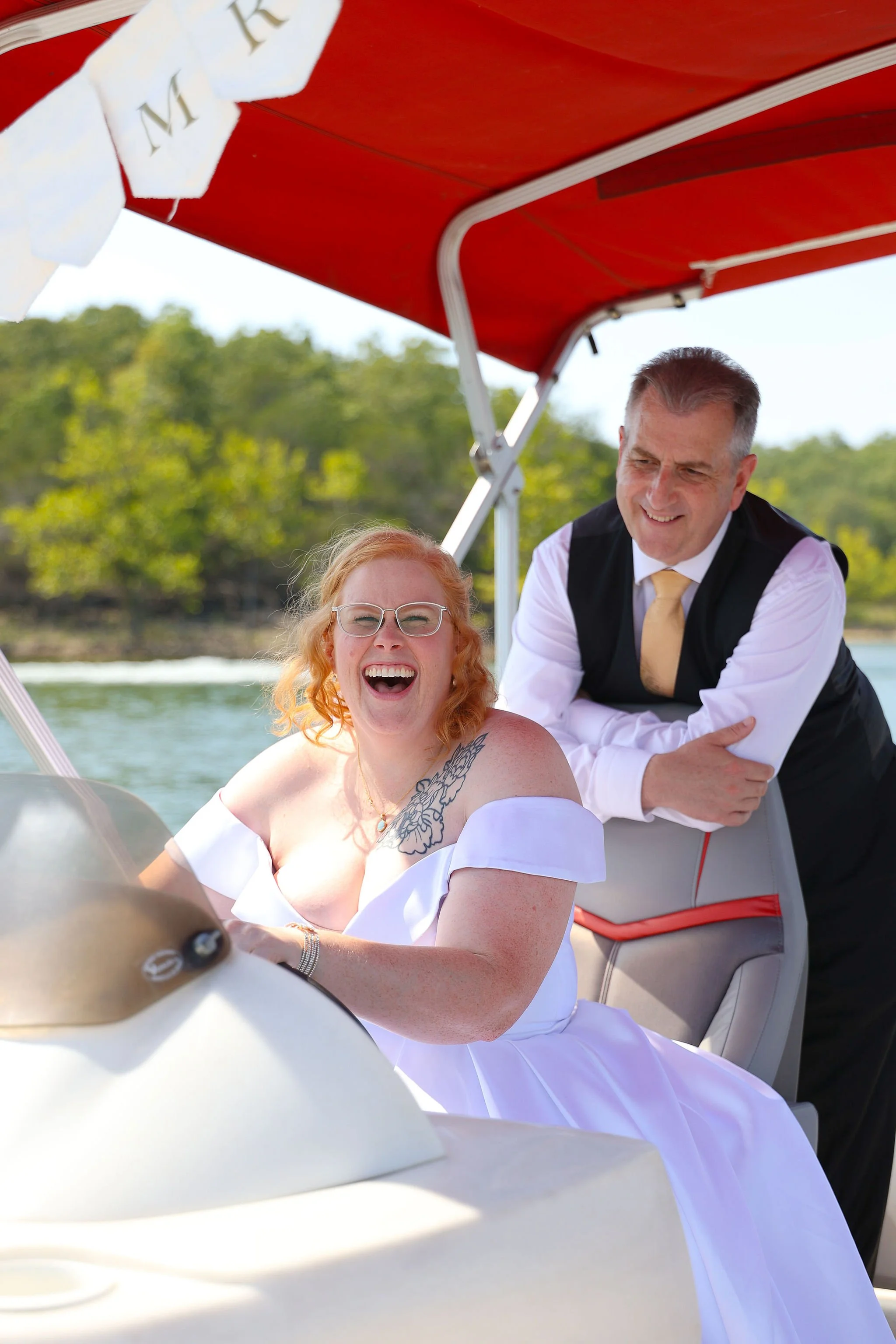 A woman with red hair, glasses, and a white off-shoulder dress is sitting on a boat, smiling and laughing, with a man standing behind her, dressed in a white shirt, black vest, and yellow tie. They are under a red canopy with a white pennant banner that has the letter 'M' on it, and the background shows water and green trees.