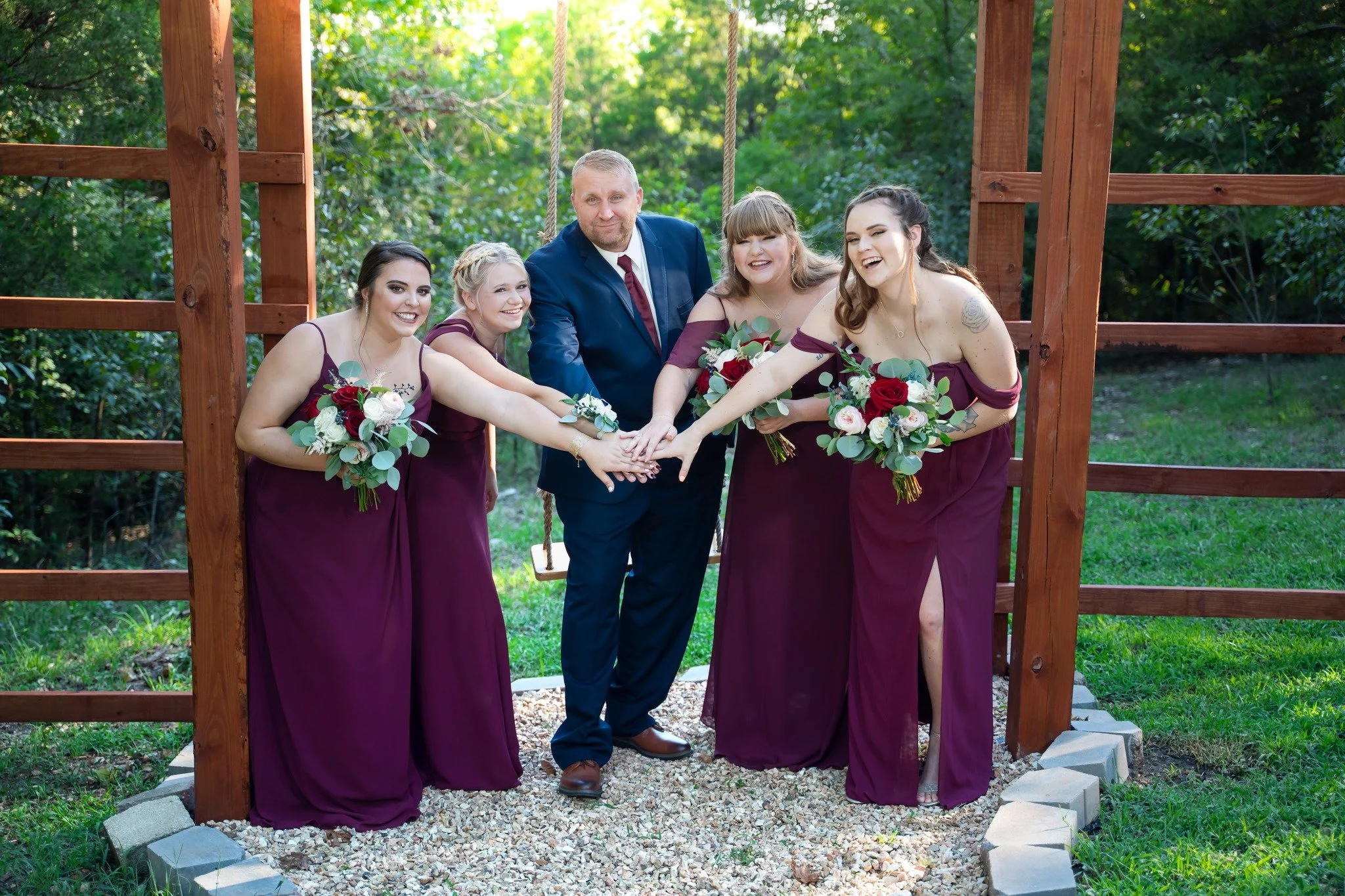 Group of six people, five women in matching purple dresses and one man in a suit, standing outdoors and holding hands. They are smiling and holding bouquets of flowers, with a wooden swing set and green trees in the background.