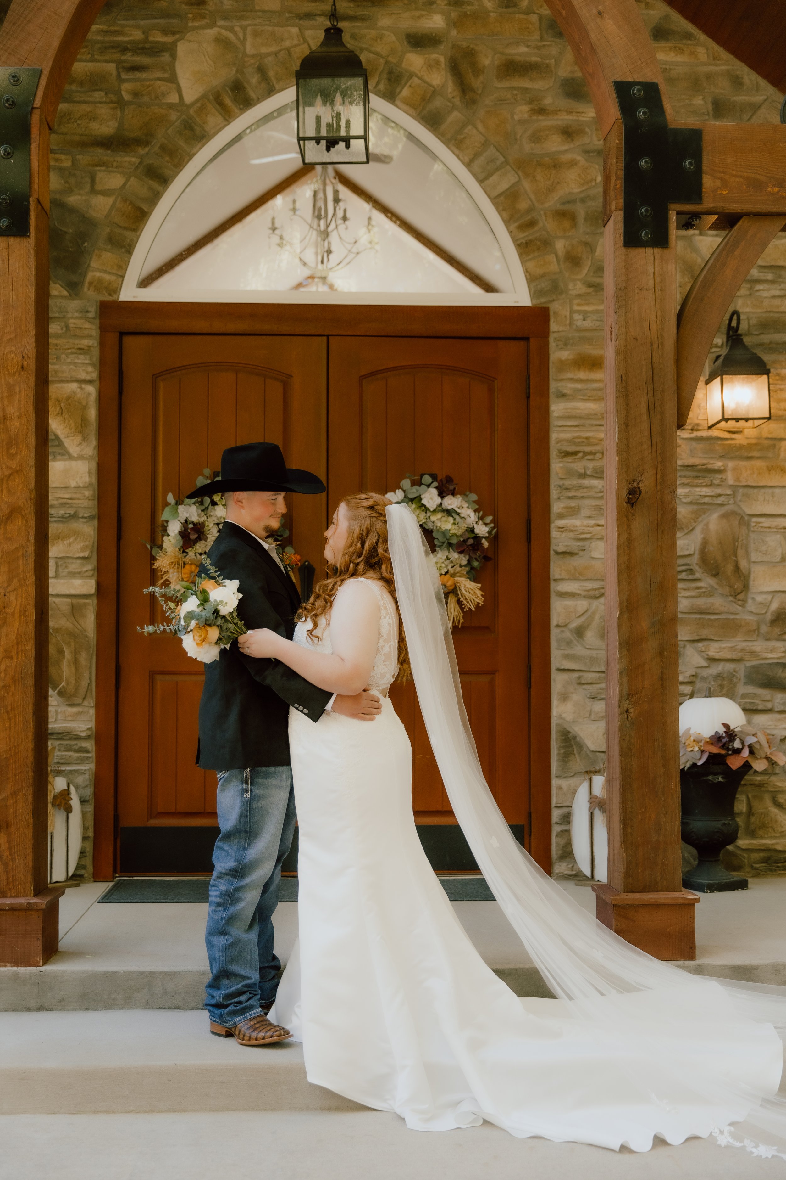 A bride and groom standing close together indoors, holding each other's hands, with floral arrangements and wooden doors in the background.