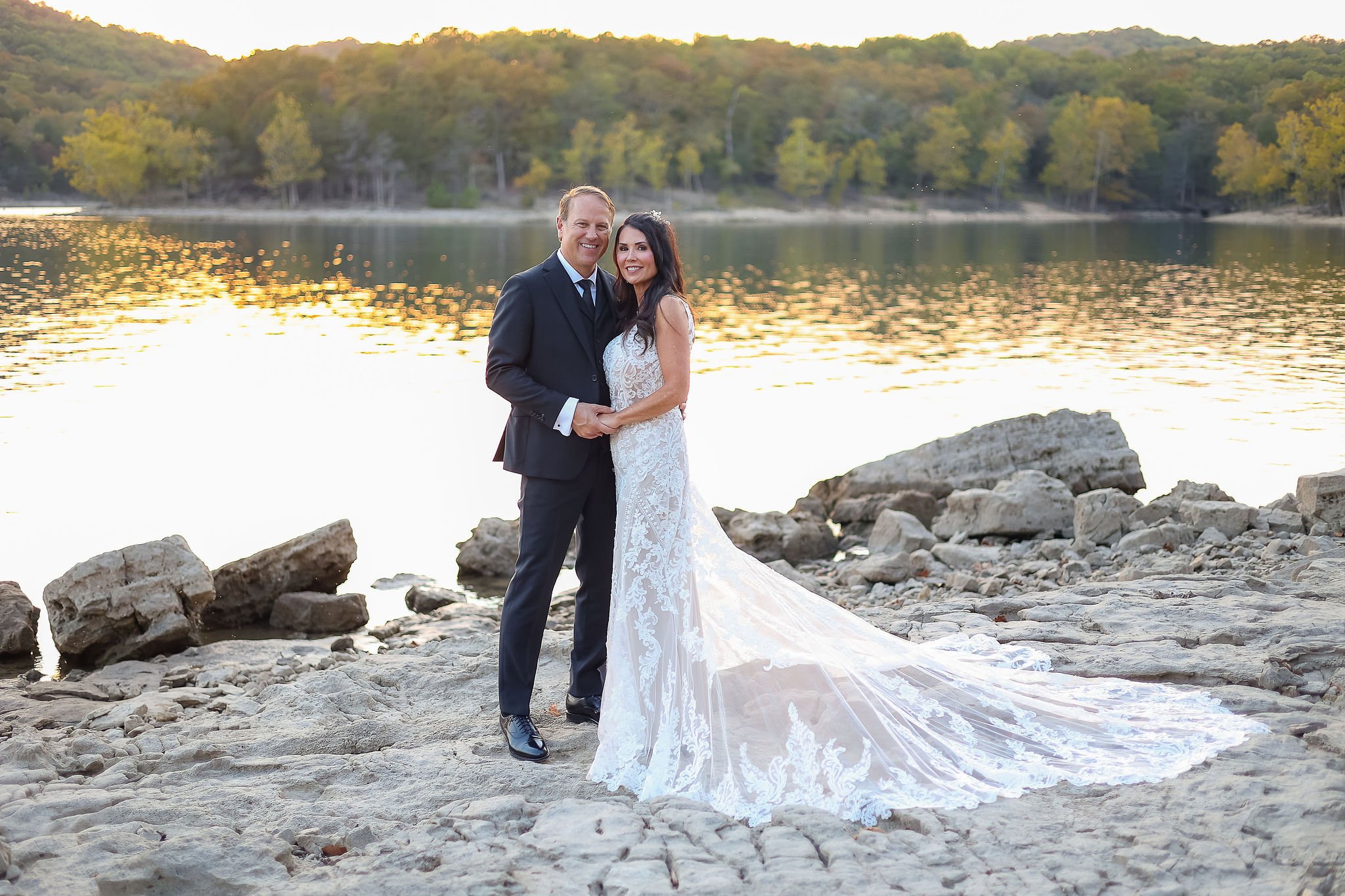 A newlywed couple stands on a rocky riverside during sunset, smiling and holding hands, with a backdrop of water, trees, and distant hills.