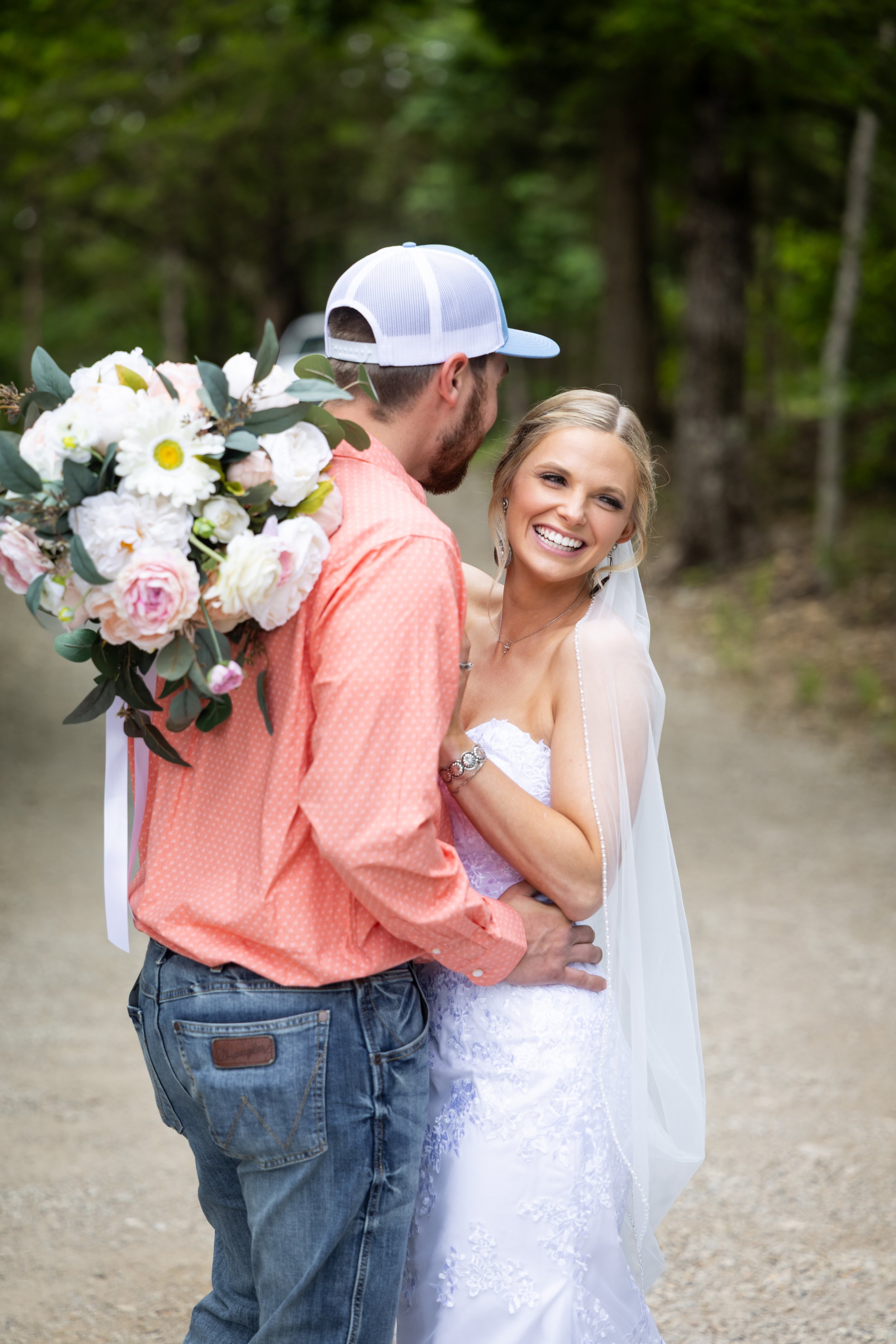 A happy bride and groom embrace outdoors, with the groom holding a bouquet of white and pink flowers, surrounded by green trees.