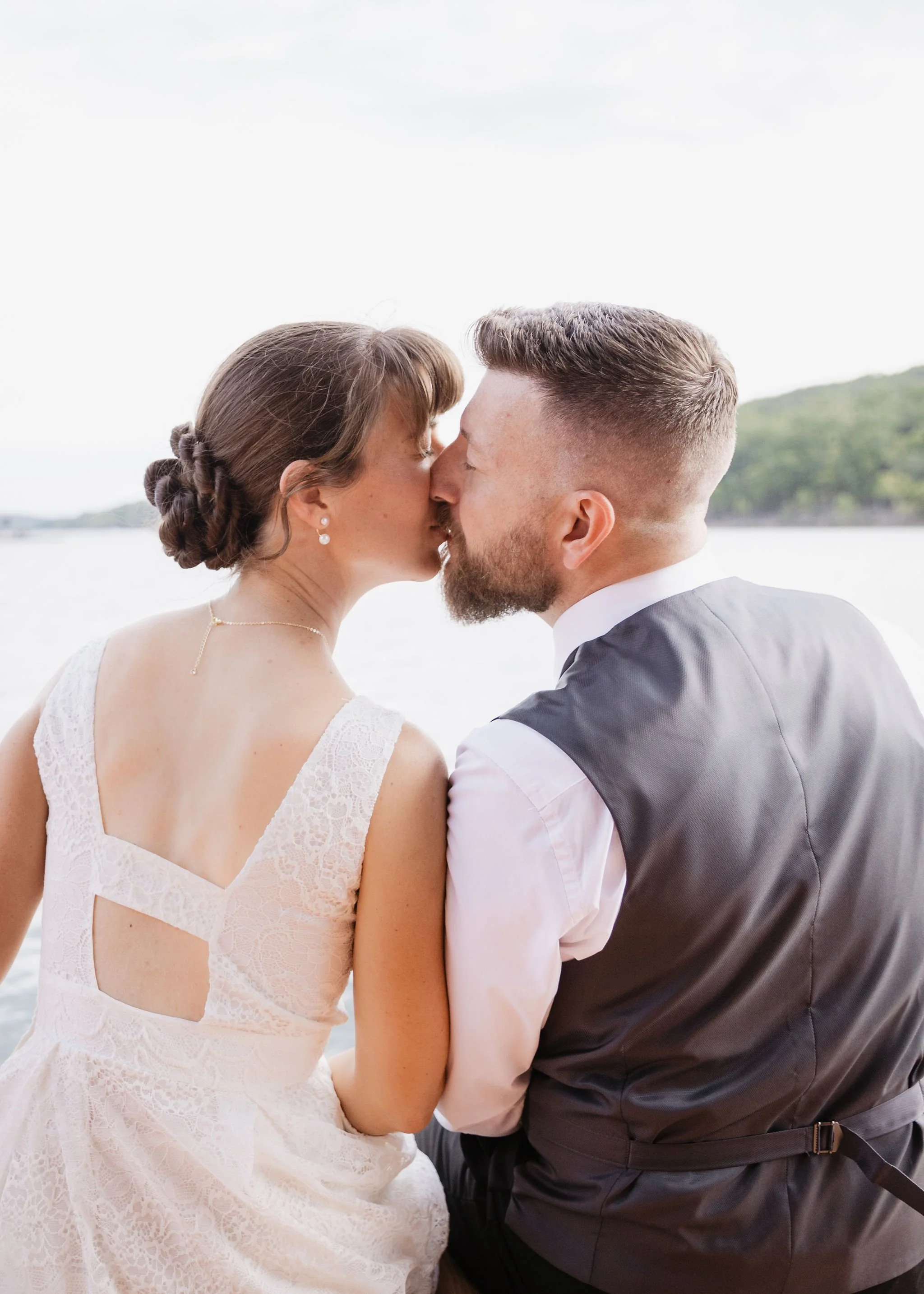 A bride and groom sharing a kiss outdoors by a lake, with the bride wearing a lace wedding dress and the groom in a gray vest and white shirt.