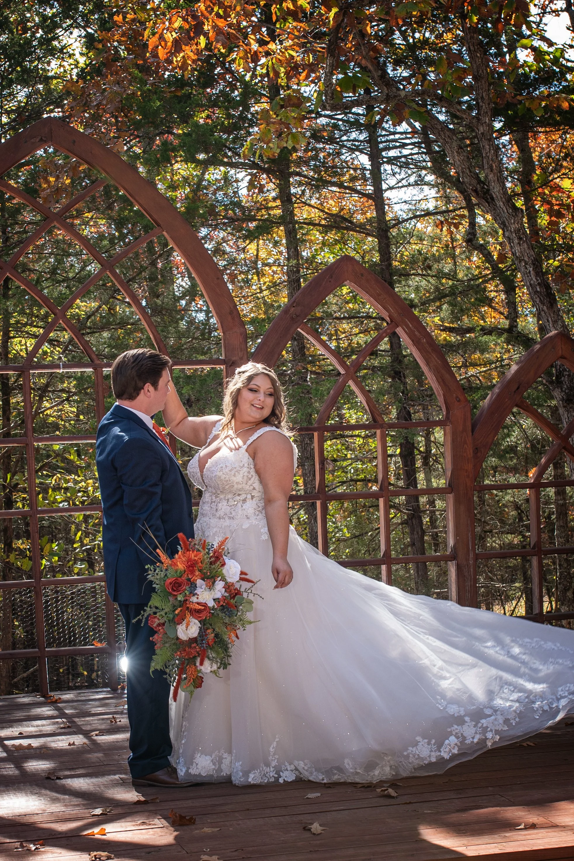 A bride and groom dancing outdoors on a wooden deck surrounded by trees with autumn leaves. The bride wears a white lace wedding gown and holds a bouquet of orange, white, and green flowers. The groom wears a dark suit and looks at the bride, who is smiling.