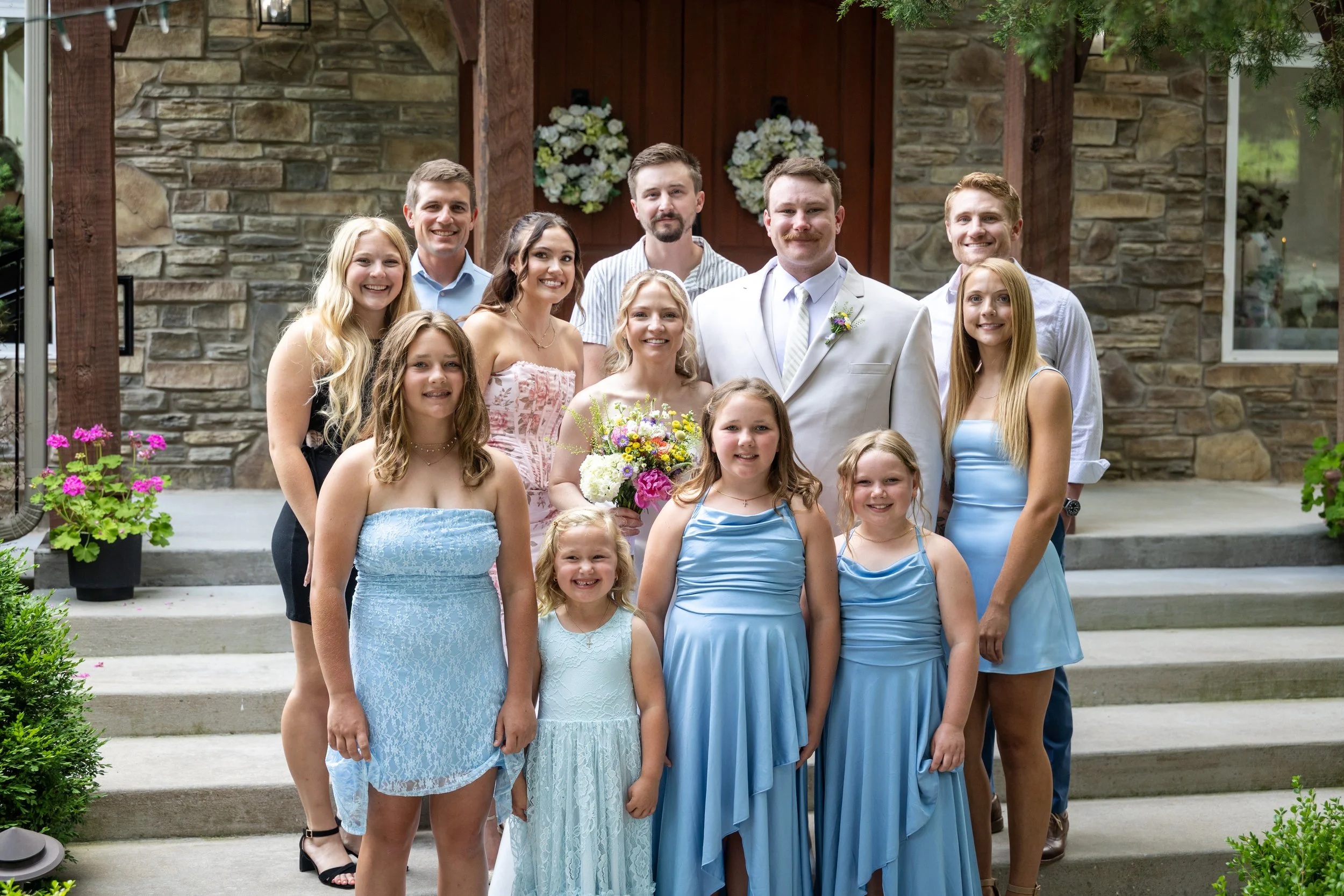 A group of people, including children and adults, dressed in formal attire, standing on steps in front of a stone house with floral decorations, celebrating a special occasion.