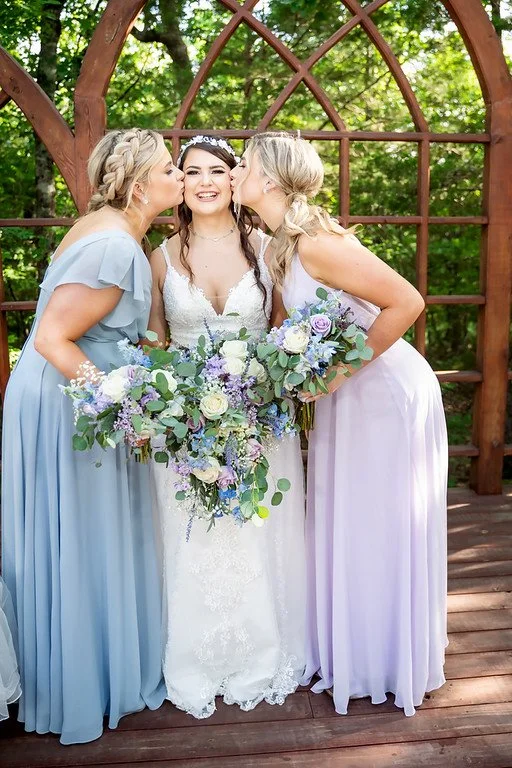 A bride with two bridesmaids kissing her on each cheek during a wedding portrait, outdoors with green trees and wooden structures in the background.