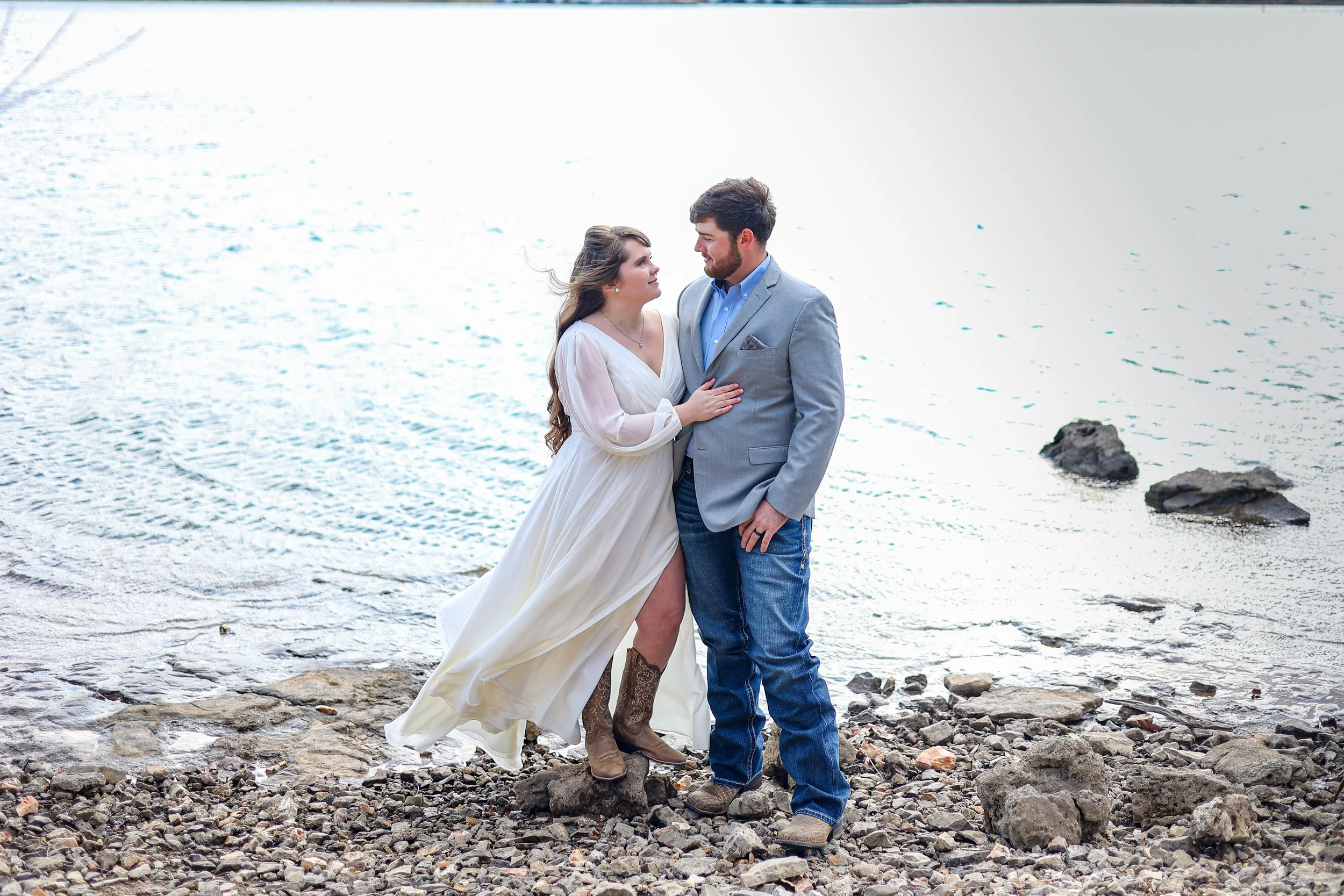 A couple standing on a rocky beach by the water, gazing at each other, with the woman wearing a white dress and the man in a gray blazer and jeans.