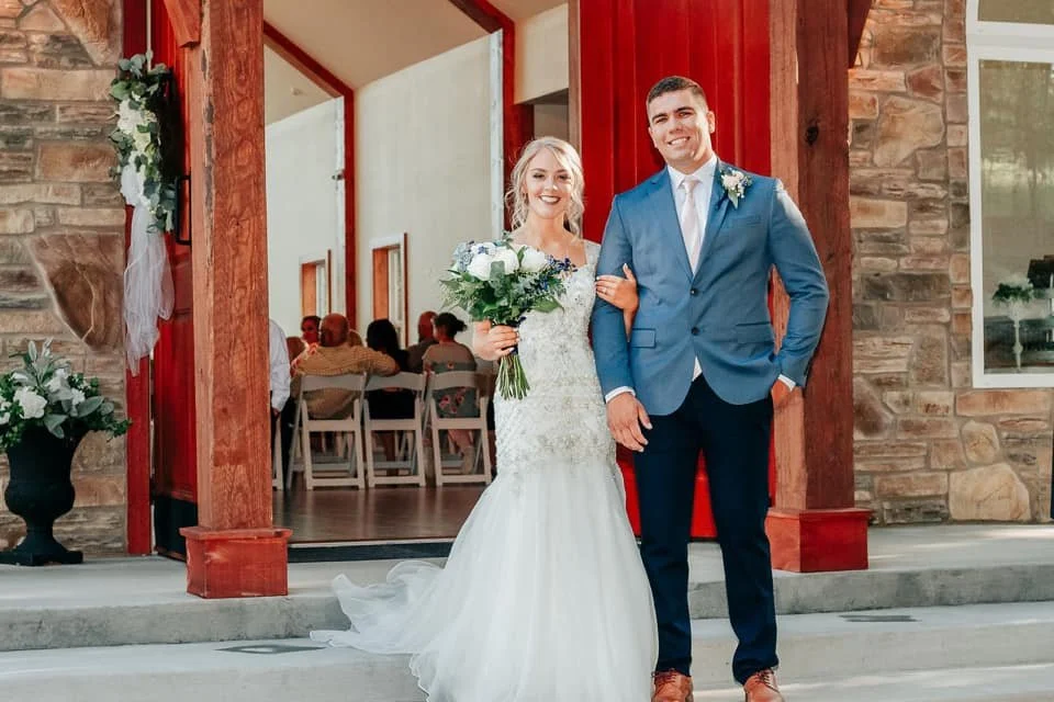 A bride in a white wedding gown holding a bouquet of white and blue flowers and a groom in a blue suit with a white shirt and tie, standing together smiling outside a building with wooden beams and stone walls, during a wedding celebration.