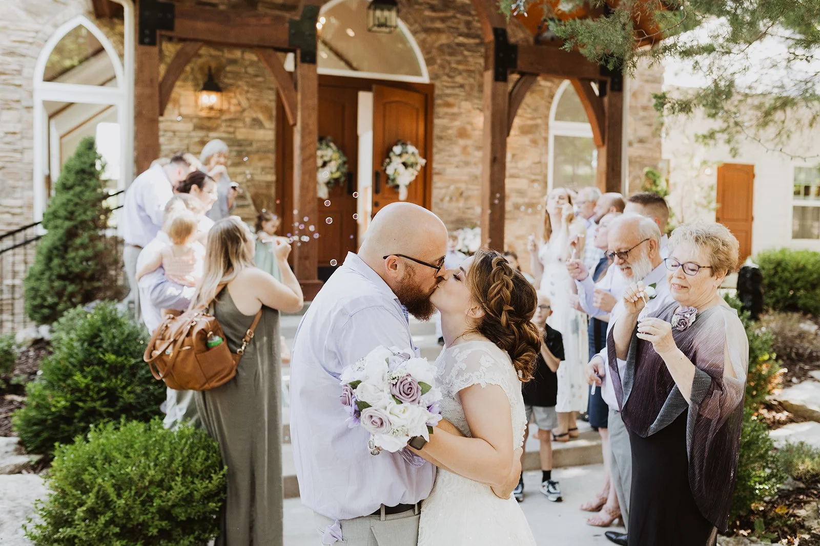 A newlywed couple sharing a kiss outside a church, holding a bouquet of flowers, surrounded by family and friends during a wedding celebration.