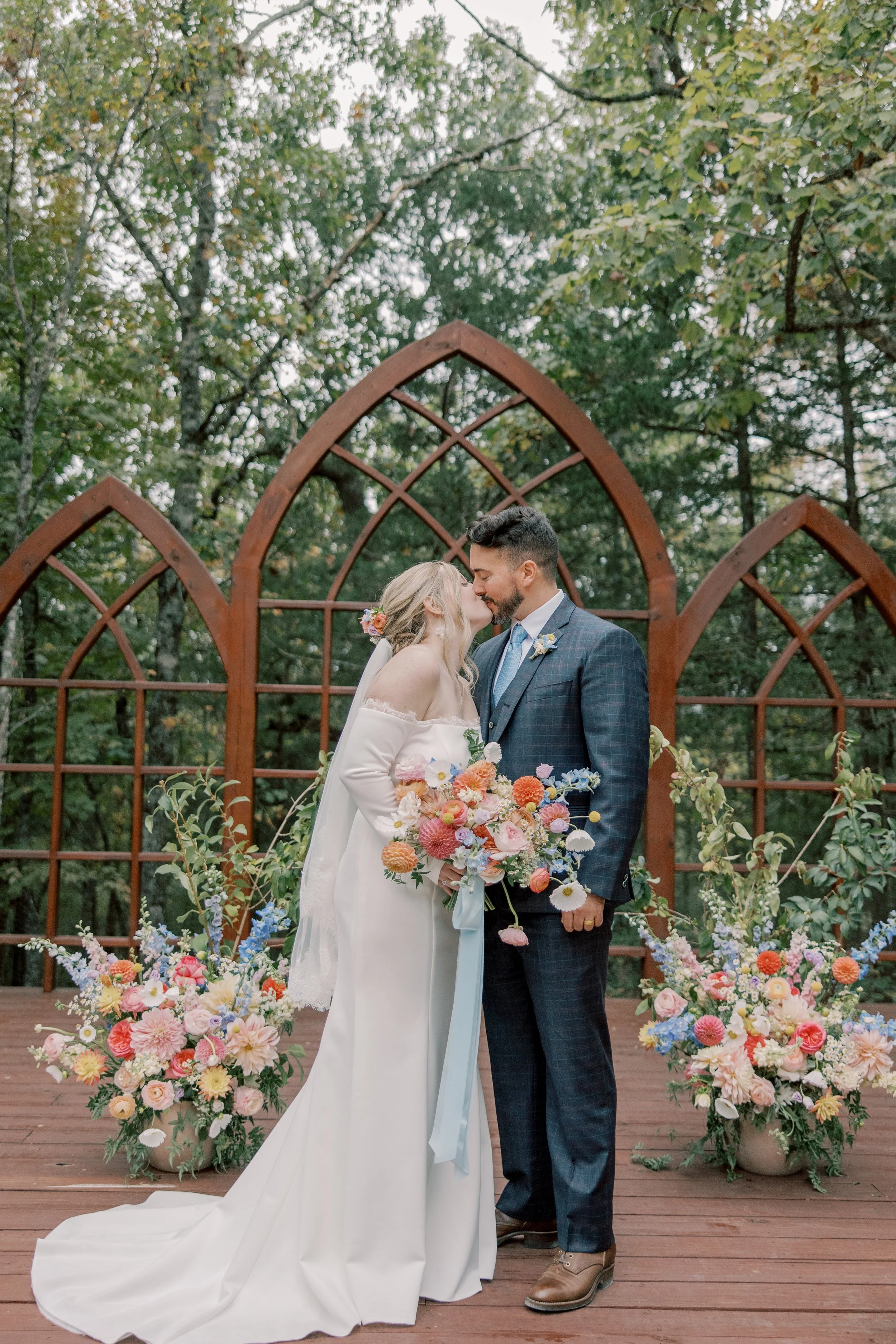 A bride and groom sharing a kiss at their wedding outdoors, surrounded by flowers and a wooden arch.