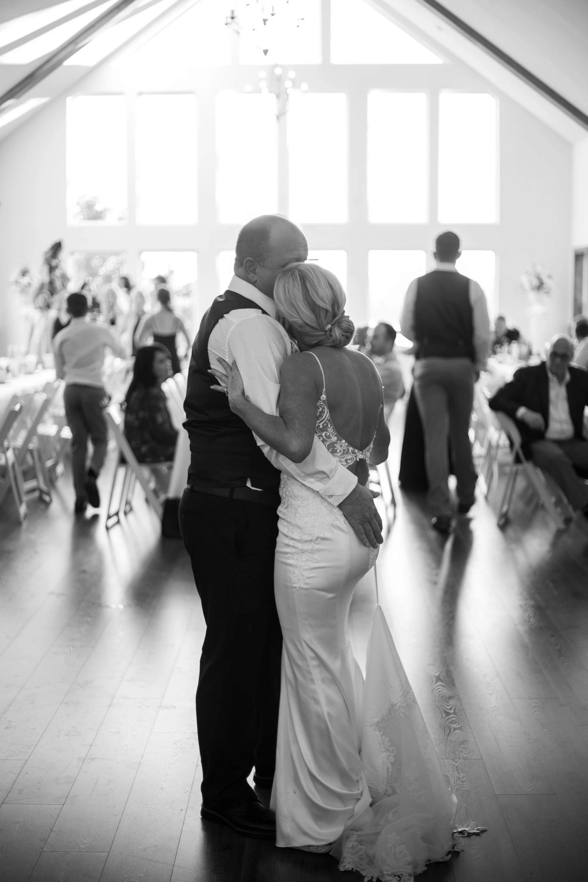 A couple dancing closely at their wedding reception in a brightly lit hall, with guests seated and standing around.