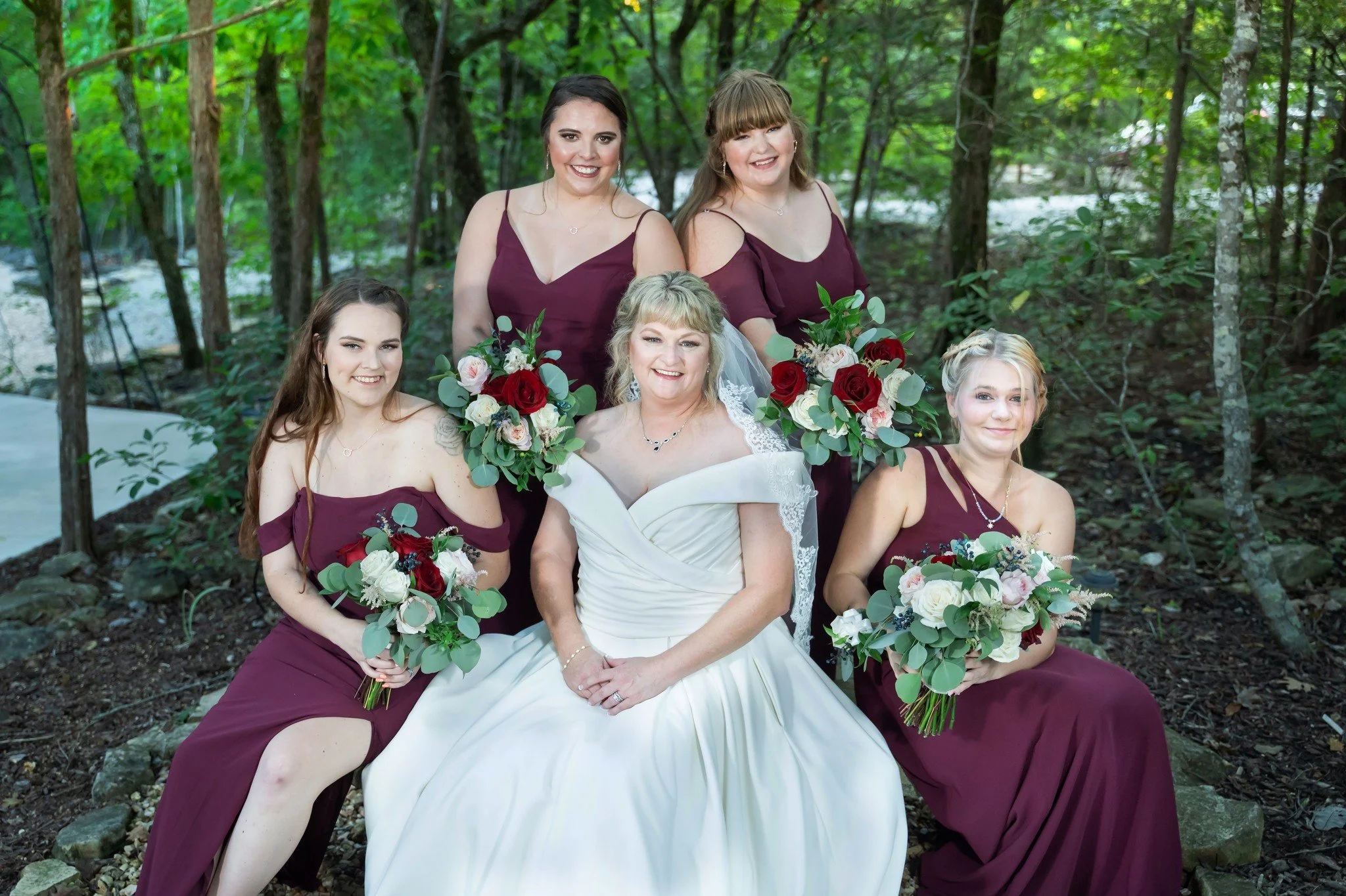 A group of six women, including a bride in a white wedding dress, holding bouquets, posing for a photo outdoors in a wooded area, all smiling.