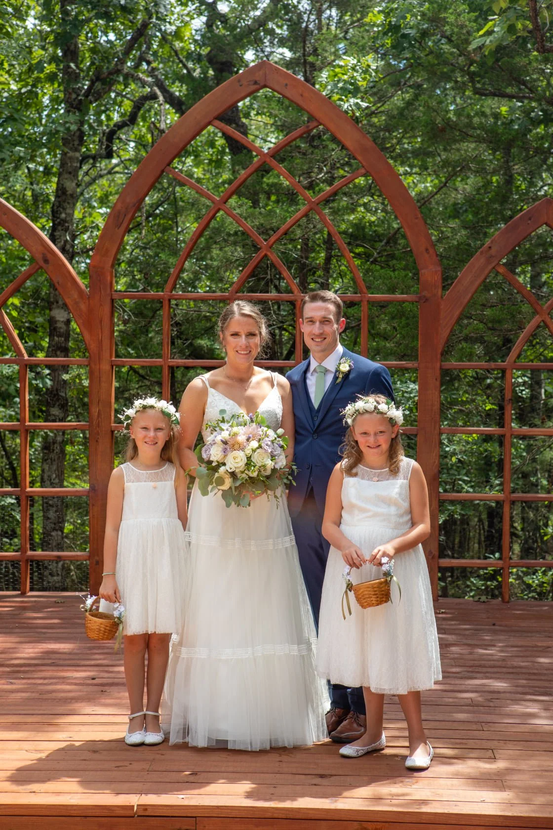 A bride and groom standing with two young girls at an outdoor wedding. The bride holds a bouquet, and the girls hold baskets, all dressed in white, with a wooden decorative backdrop and green trees in the background.