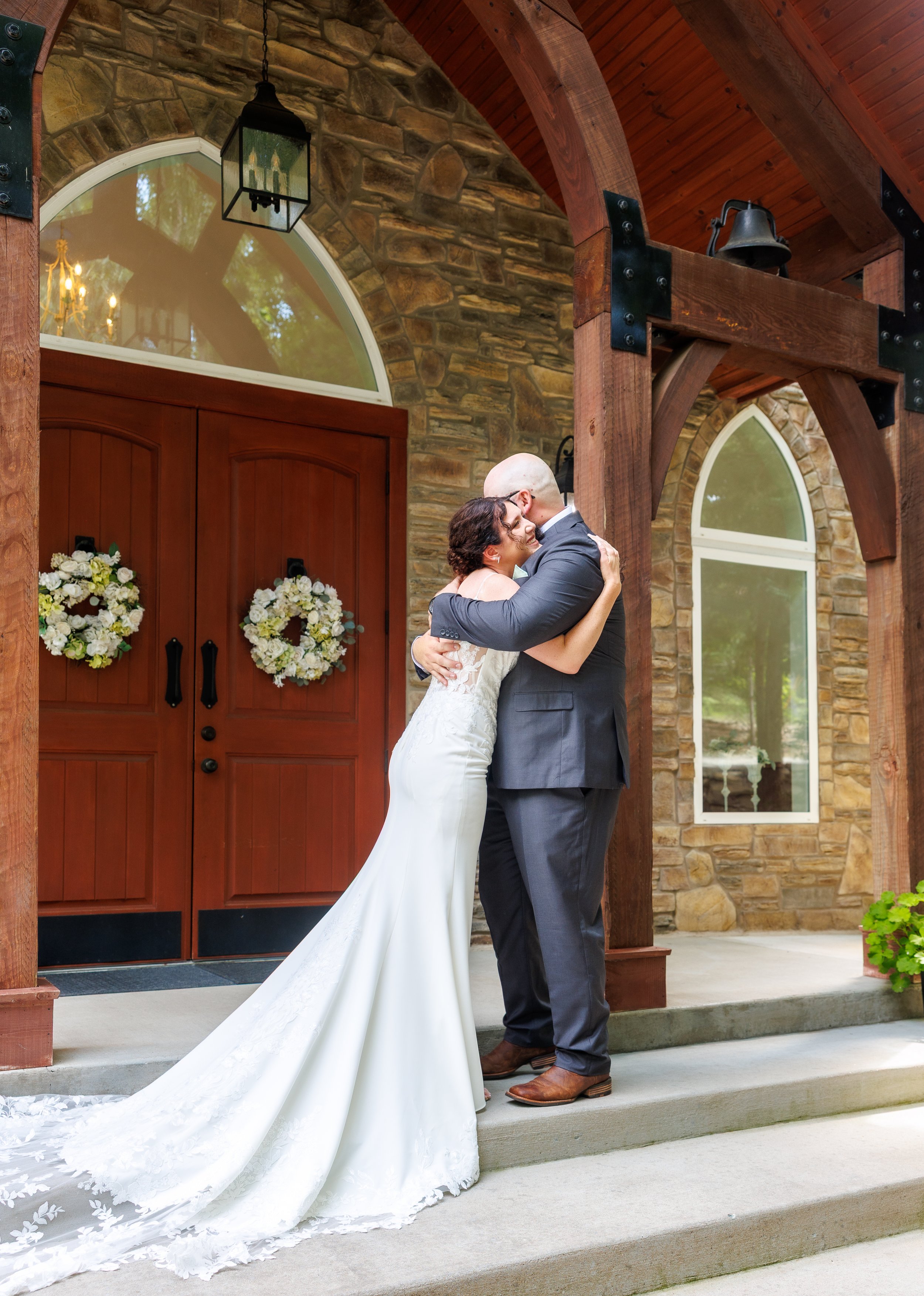A bride and groom hugging on the steps outside a stone and wood building with large arched windows and decorated with floral wreaths on the doors.