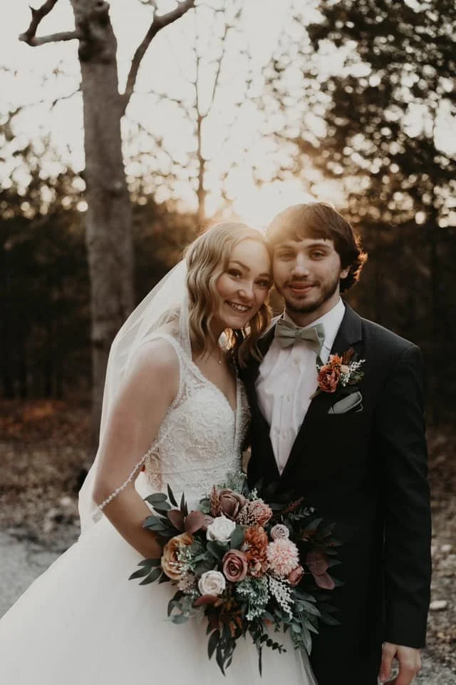 A bride and groom posing outdoors at sunset. The bride is holding a bouquet of roses and greenery, wearing a lace wedding dress and veil. The groom is dressed in a black tuxedo with a bow tie and boutonniere. They are smiling and standing close toget