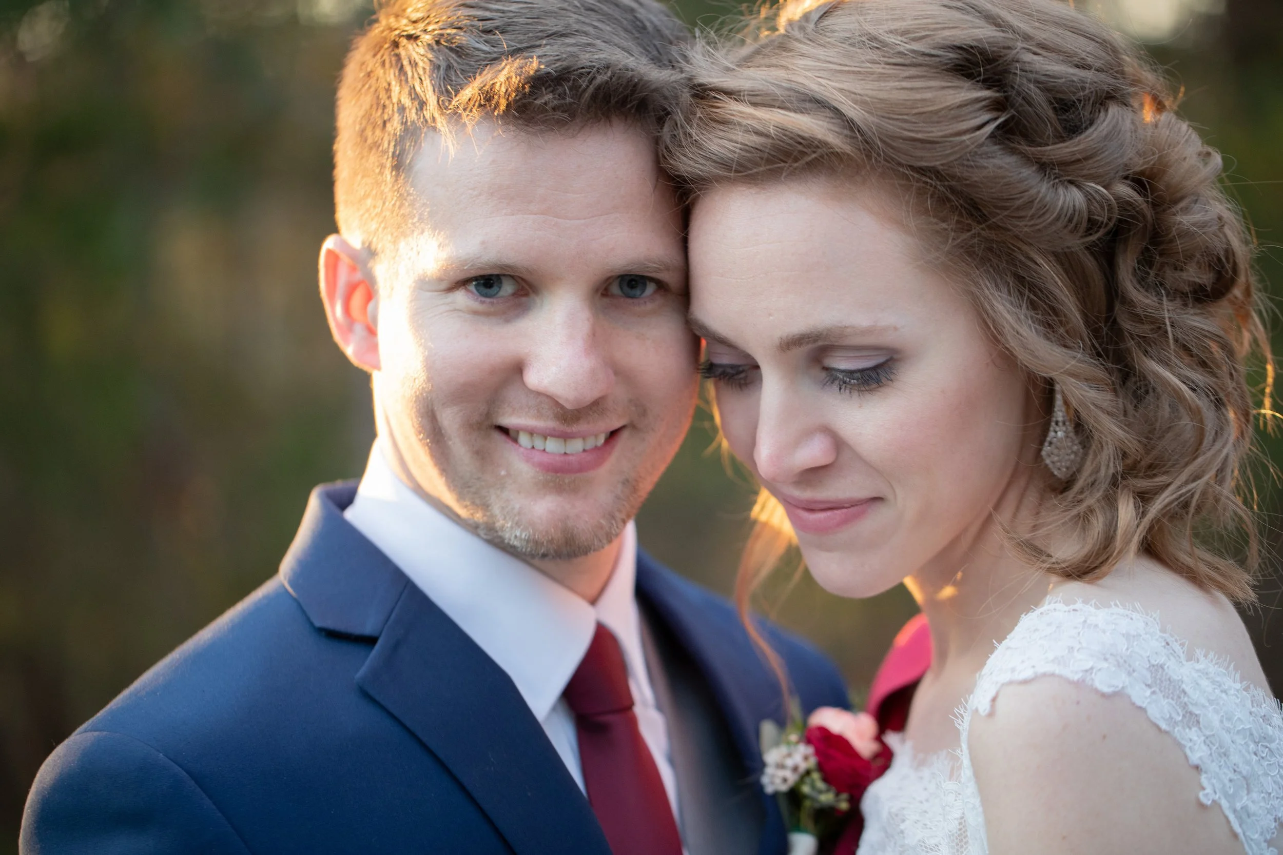 A newlywed couple, a man with blue eyes, short brown hair, and a blue suit, and a woman with curly brown hair, closed eyes, and a white lace wedding dress, embrace outdoors during sunset.