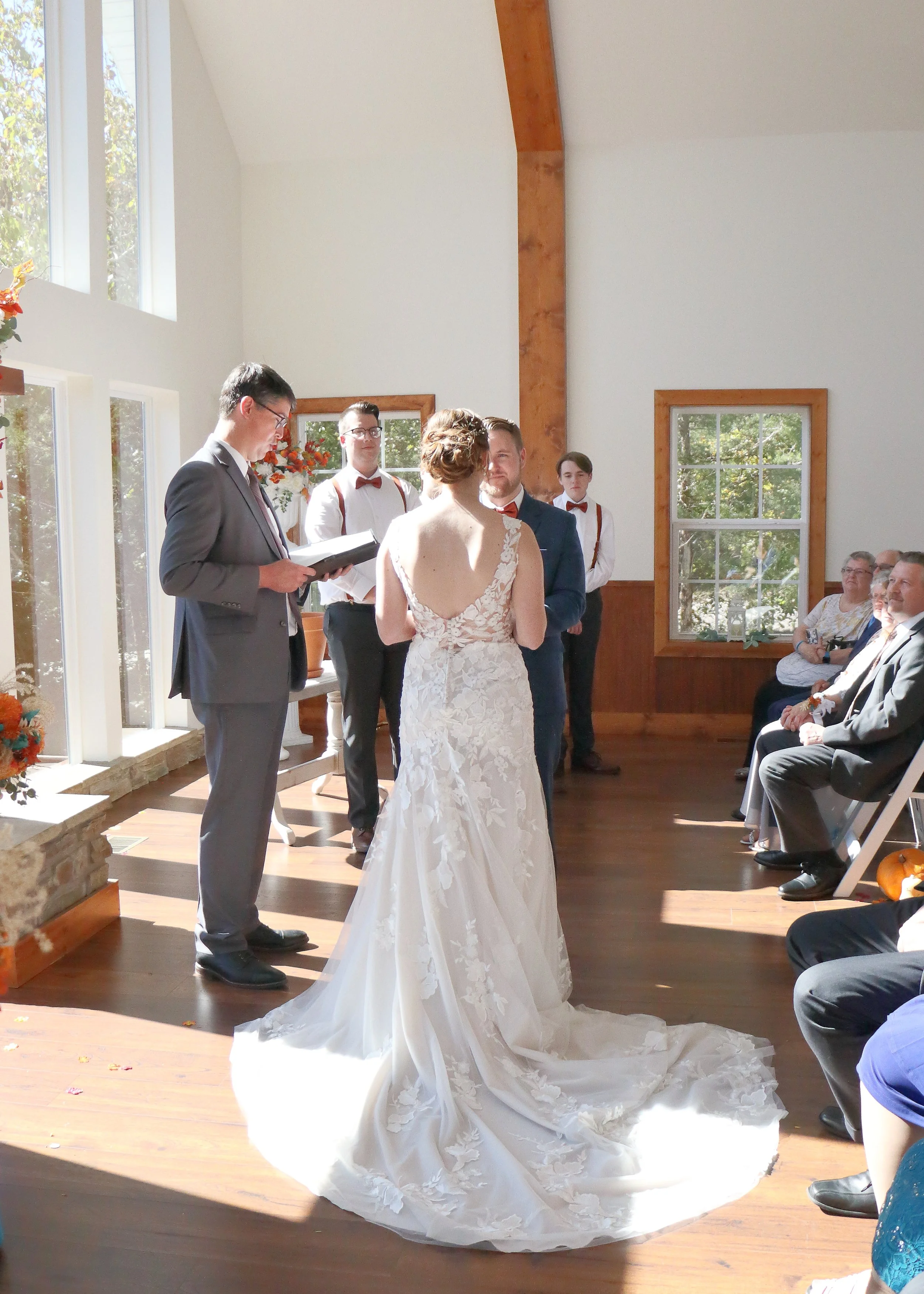 A wedding ceremony taking place indoors with a bride in a white lace gown and a groom in a blue suit exchanging vows, surrounded by guests seated on chairs, sunlight streaming through large windows, and floral decorations.