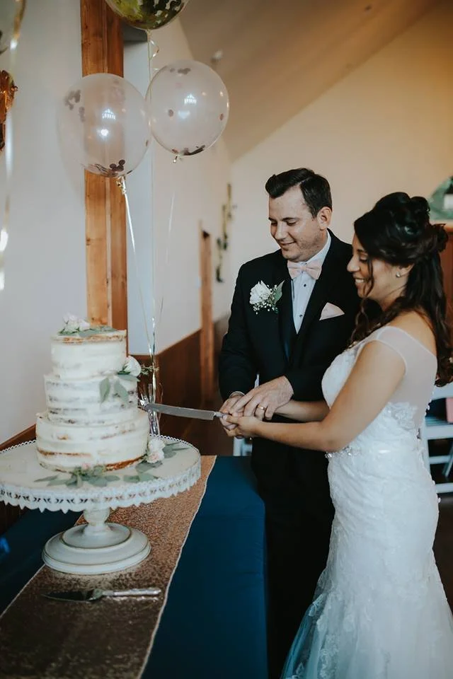 A newlywed couple cuts a wedding cake together at their celebration. The bride is wearing a white wedding gown and the groom is in a black tuxedo. There are balloons and decorations in the background.