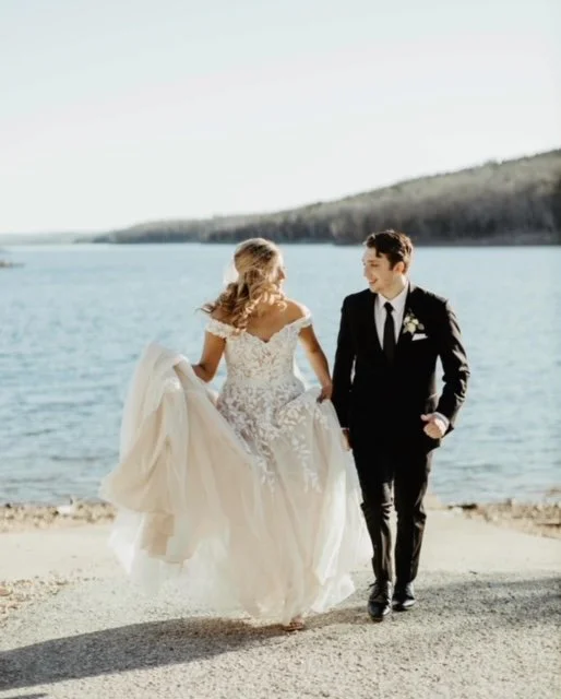 Bride and groom walking by a lake on their wedding day, holding hands, with the bride in a white gown and the groom in a black tuxedo.