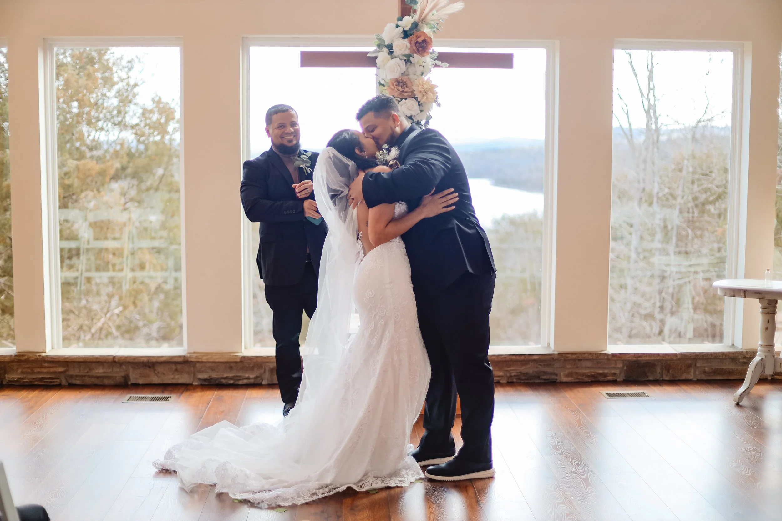 A bride and groom sharing a kiss during their wedding ceremony inside a room with large windows and a scenic outdoor landscape in the background. An officiant stands nearby smiling.