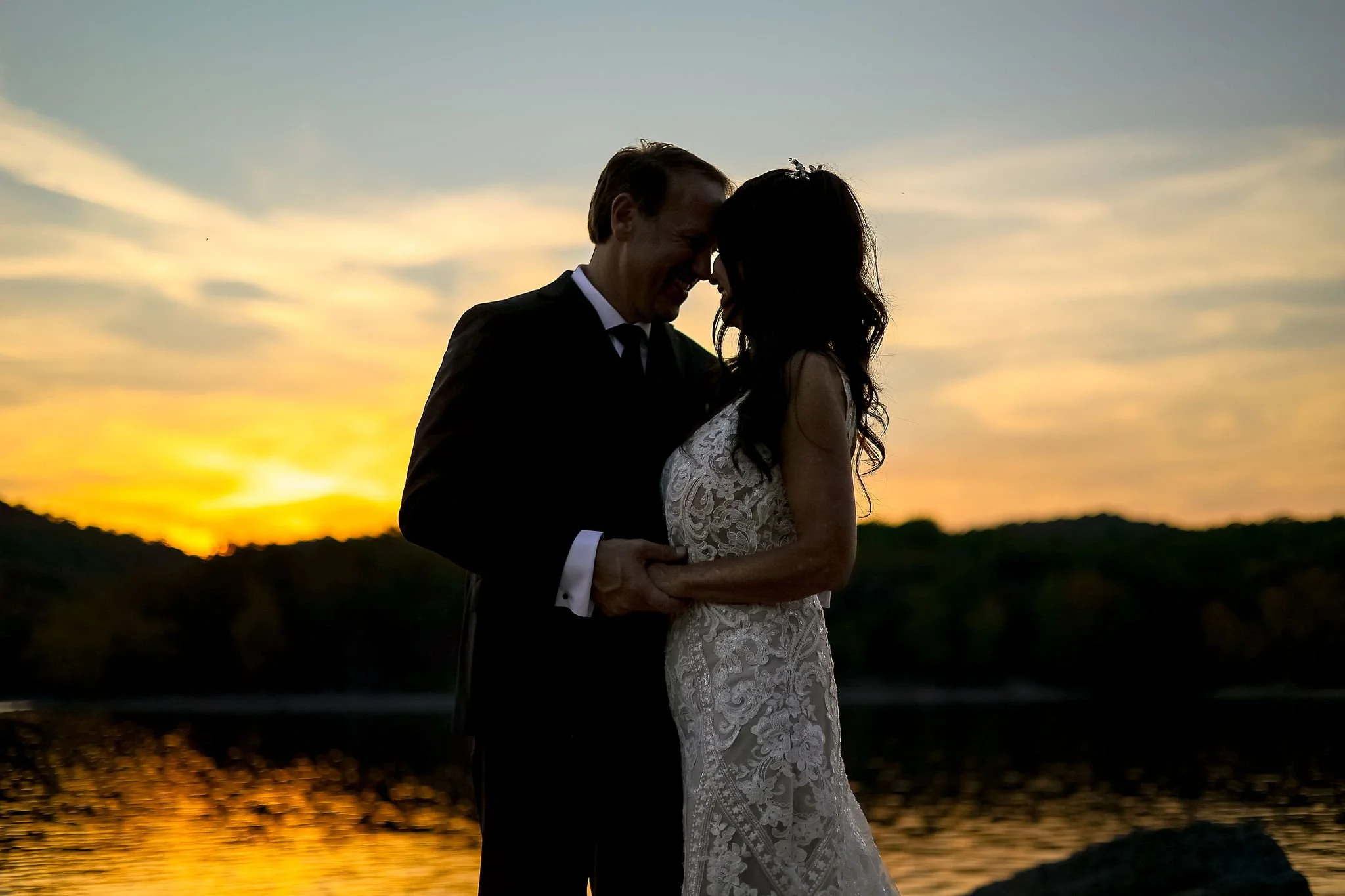 A couple in wedding attire standing close together at sunset, smiling and touching foreheads, near a body of water.