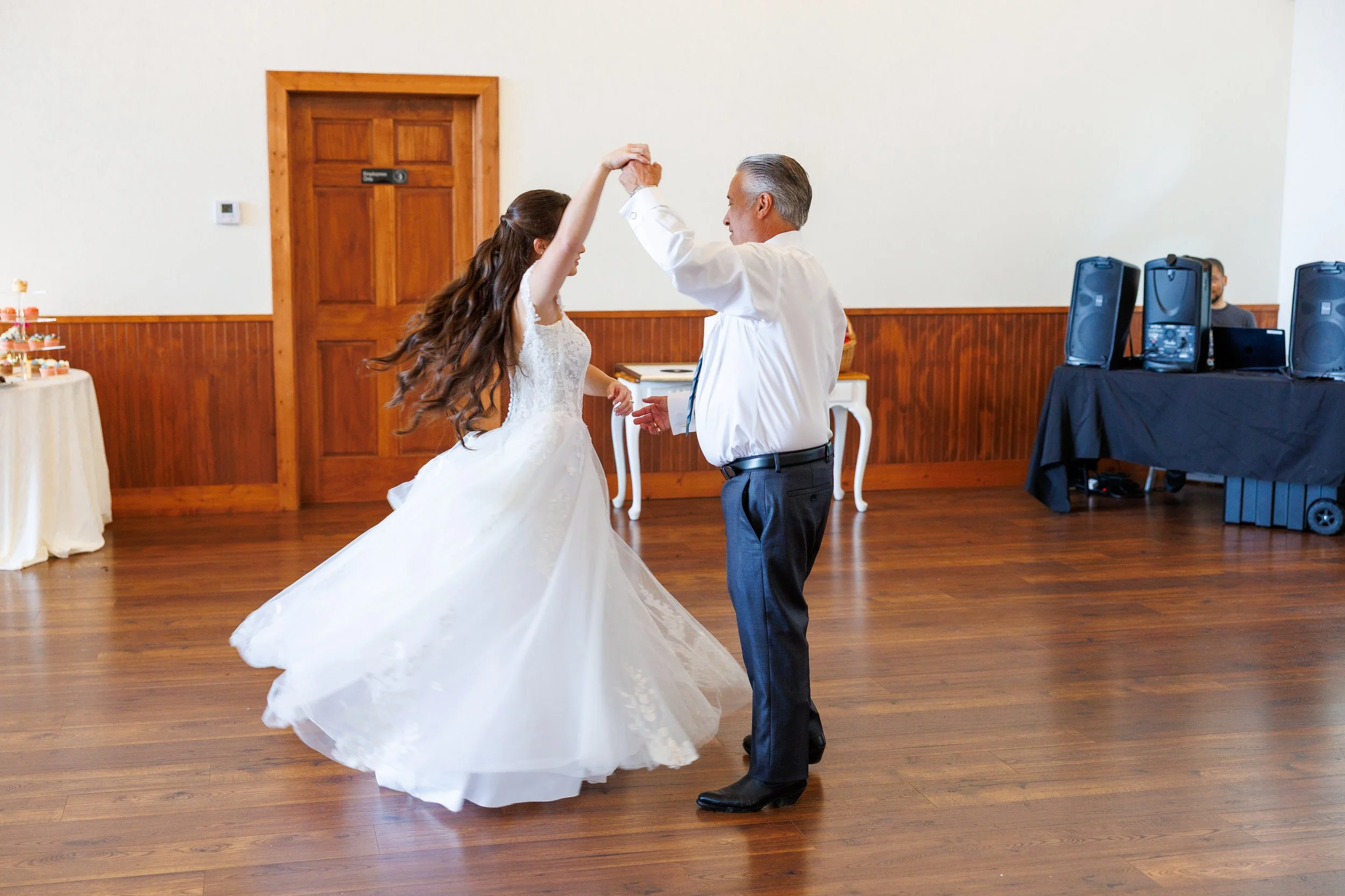 A bride and an older man dancing together in a wooden-floored reception hall. The bride is in a white wedding gown, and the man is in a white shirt and black pants.