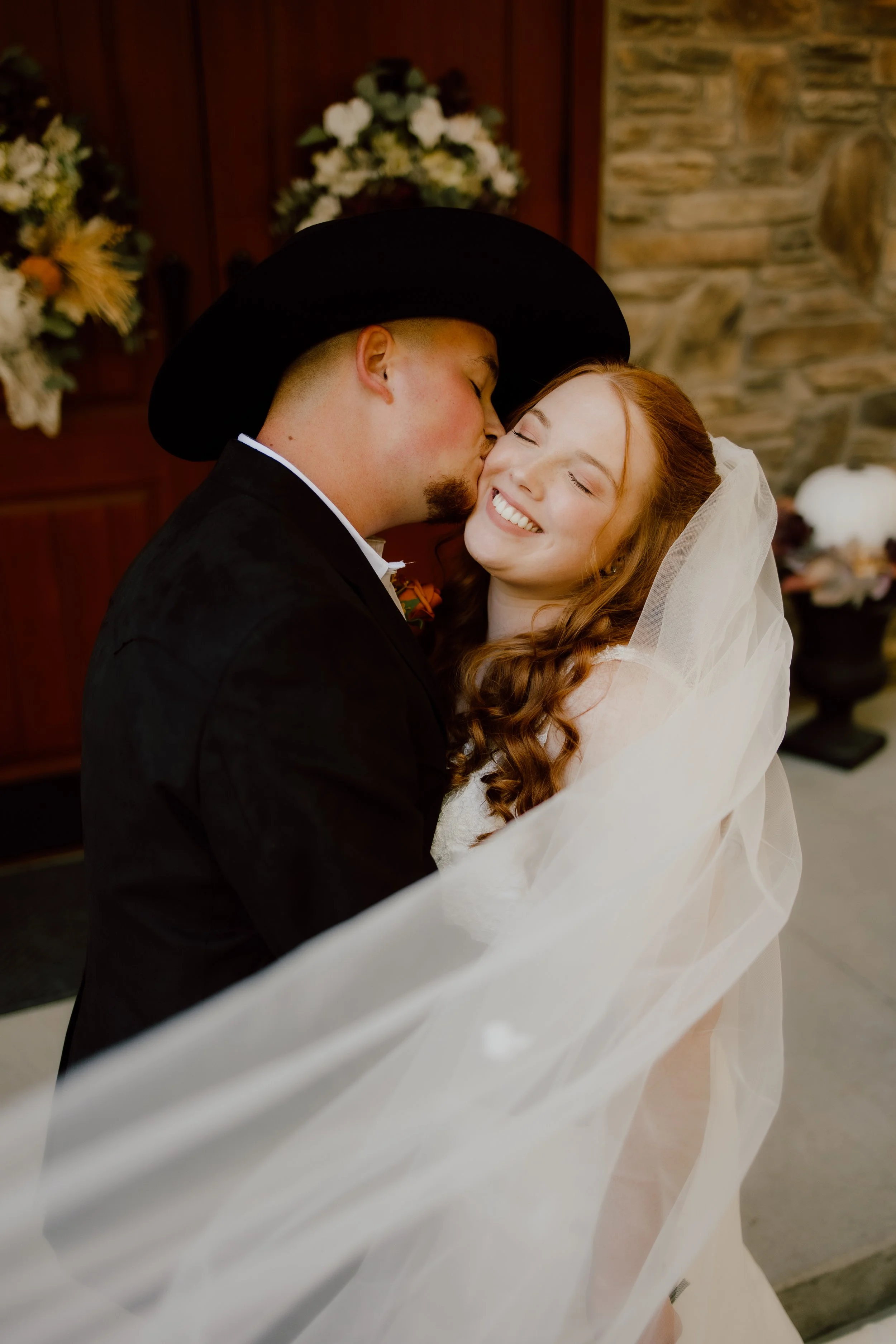 A couple dressed in wedding attire sharing a kiss, with the woman smiling and eyes closed, in an indoor setting with floral arrangements and a stone wall in the background.