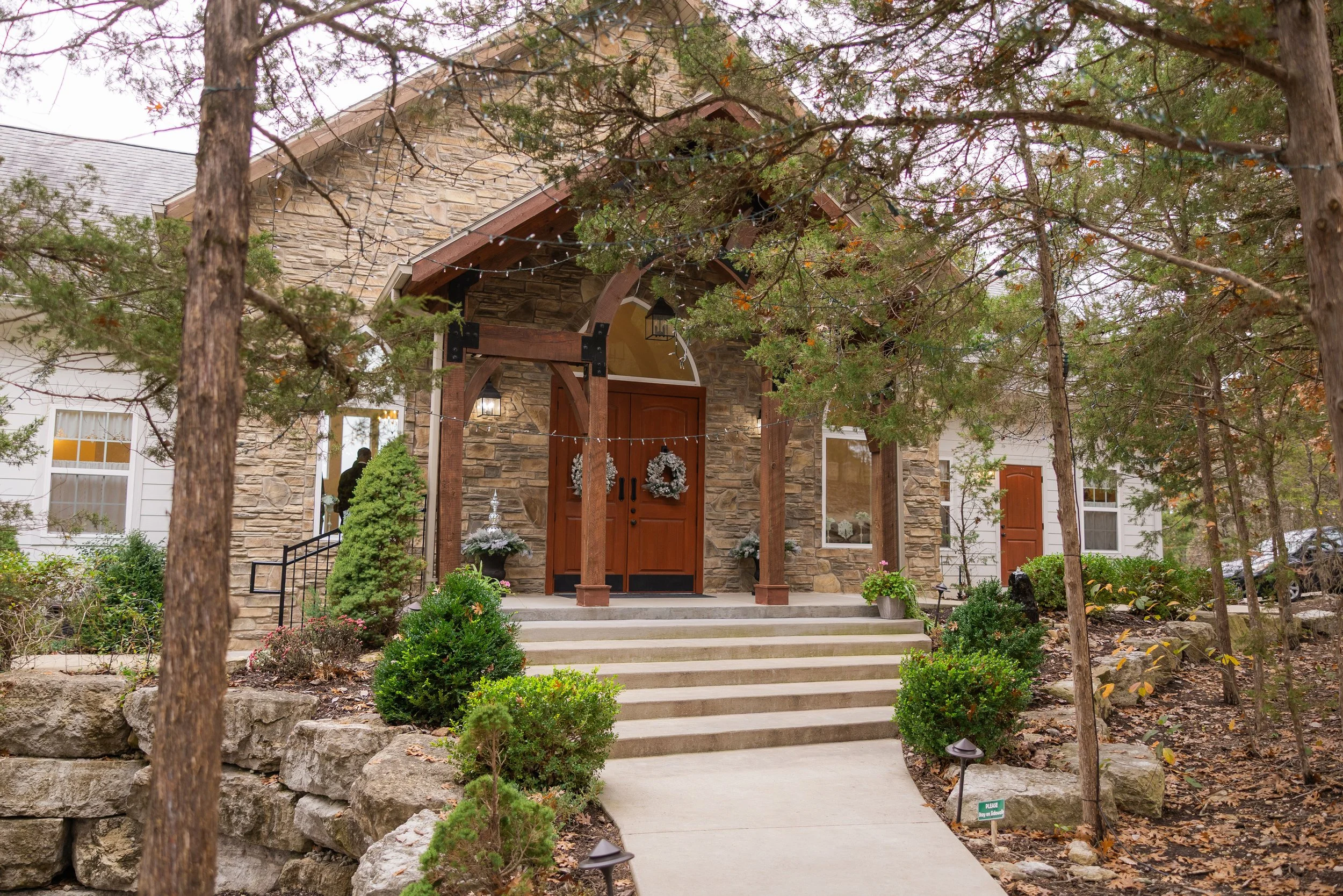 Front view of a house with a stone facade and wooden accents, partially obscured by trees and including a decorated front door with a wreath, a small porch, and steps leading up to the entrance, surrounded by landscaped greenery.