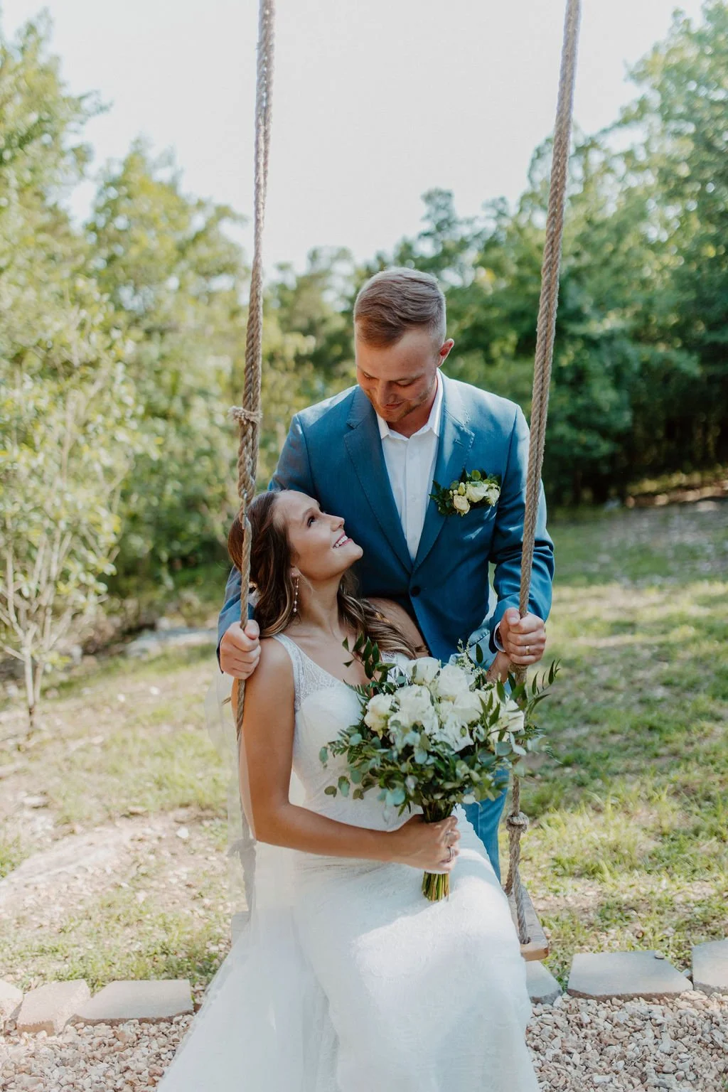 A bride sitting on a swing holding a bouquet, smiling up at a groom standing in front of her, outdoors with trees in the background.