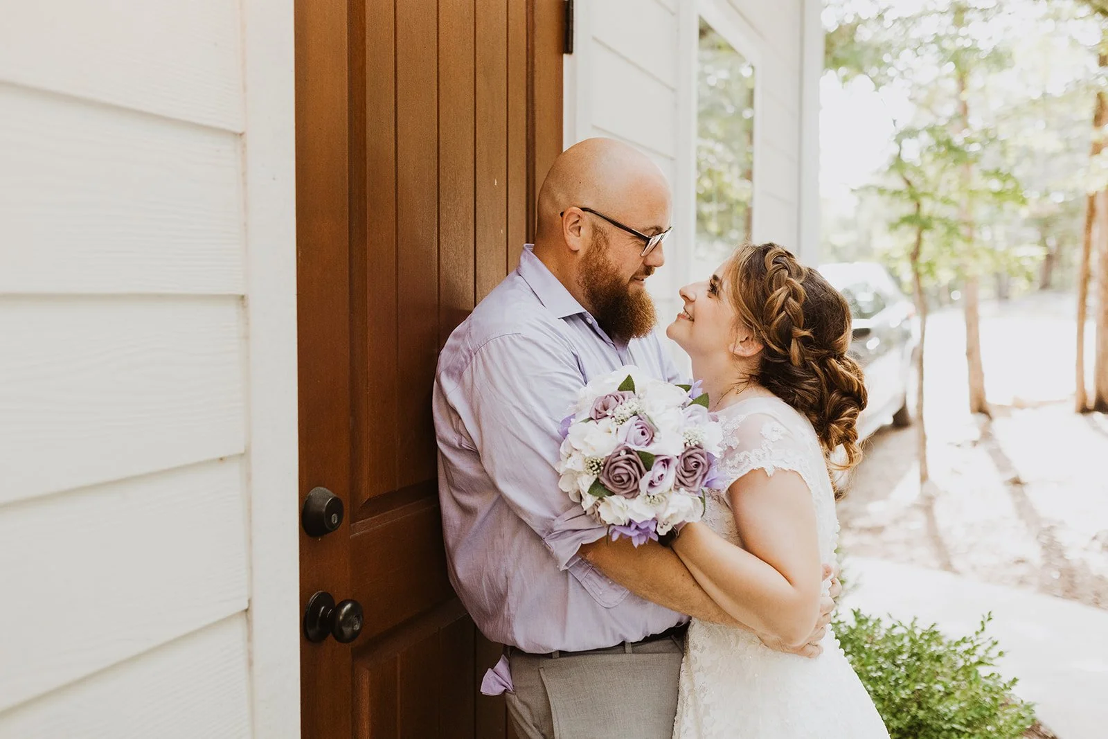 A couple sharing a romantic moment outside a house, with the man holding a bouquet of white and purple flowers, while gazing into each other's eyes.