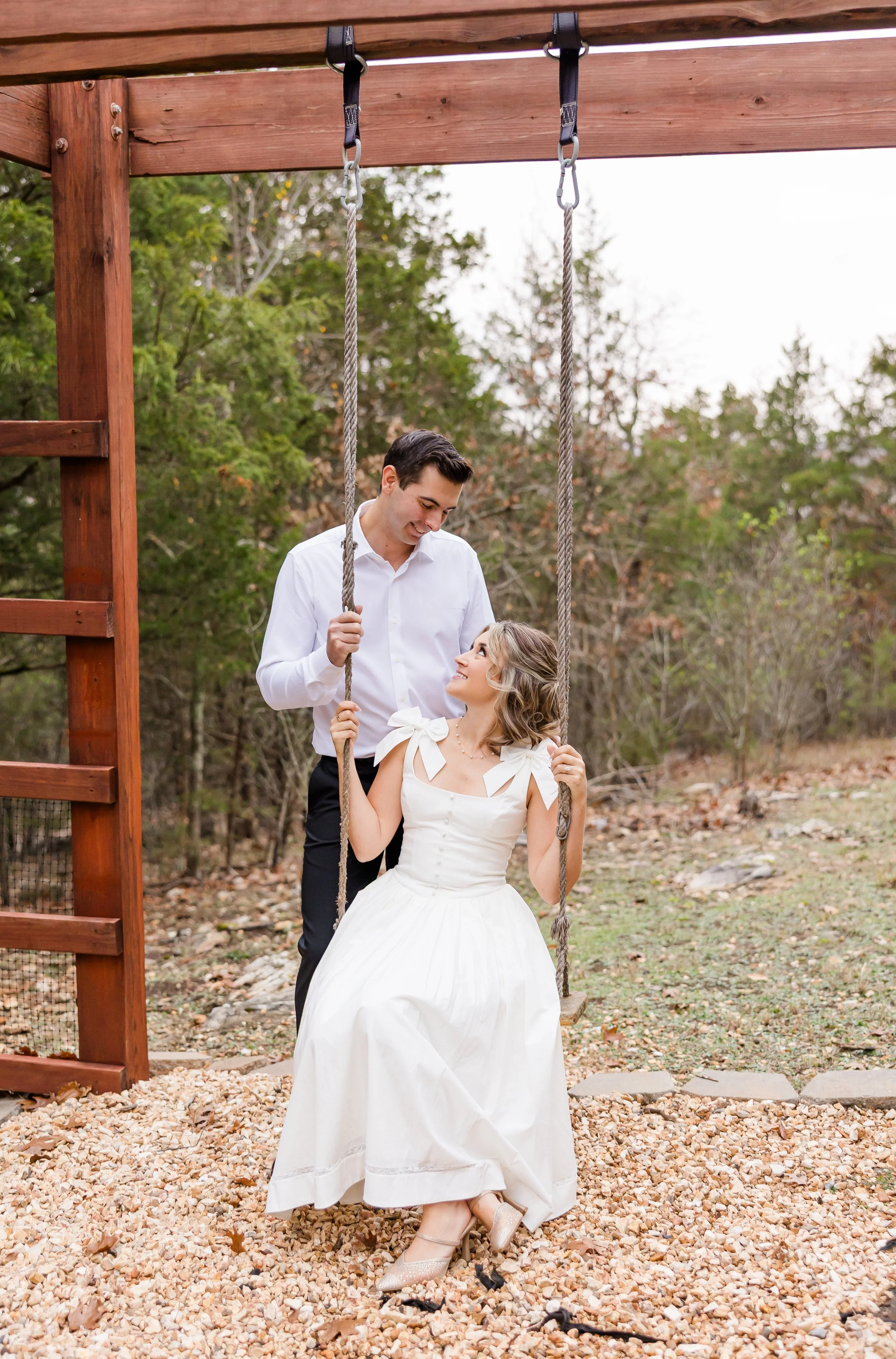 A man and woman in wedding attire on a playground swing set outdoors. The man is standing behind the woman, holding her hands, and looking at her. The woman is seated on the swing, looking up at the man, wearing a white dress with bows on the shoulders. The background has trees with fall foliage.