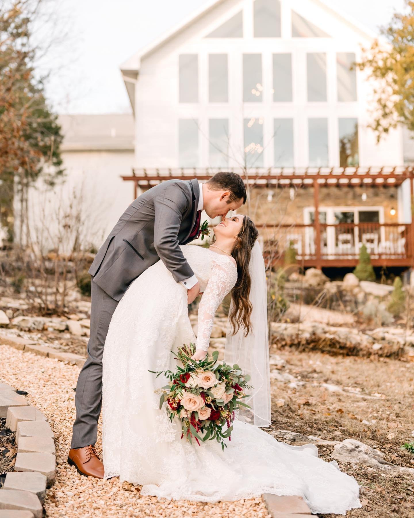 A bride and groom in wedding attire share a dip and kiss outdoors with a house in the background.