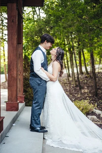 Bride and groom facing each other, holding hands, outdoors near a wooden structure with green trees in the background.