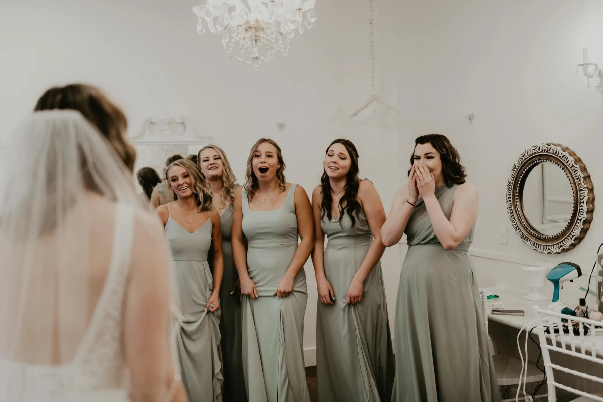 A group of women dressed in matching light green bridesmaid dresses standing together, reacting happily and excitedly in a bridal boutique or dressing room with white walls, ornate mirror, and chandelier.