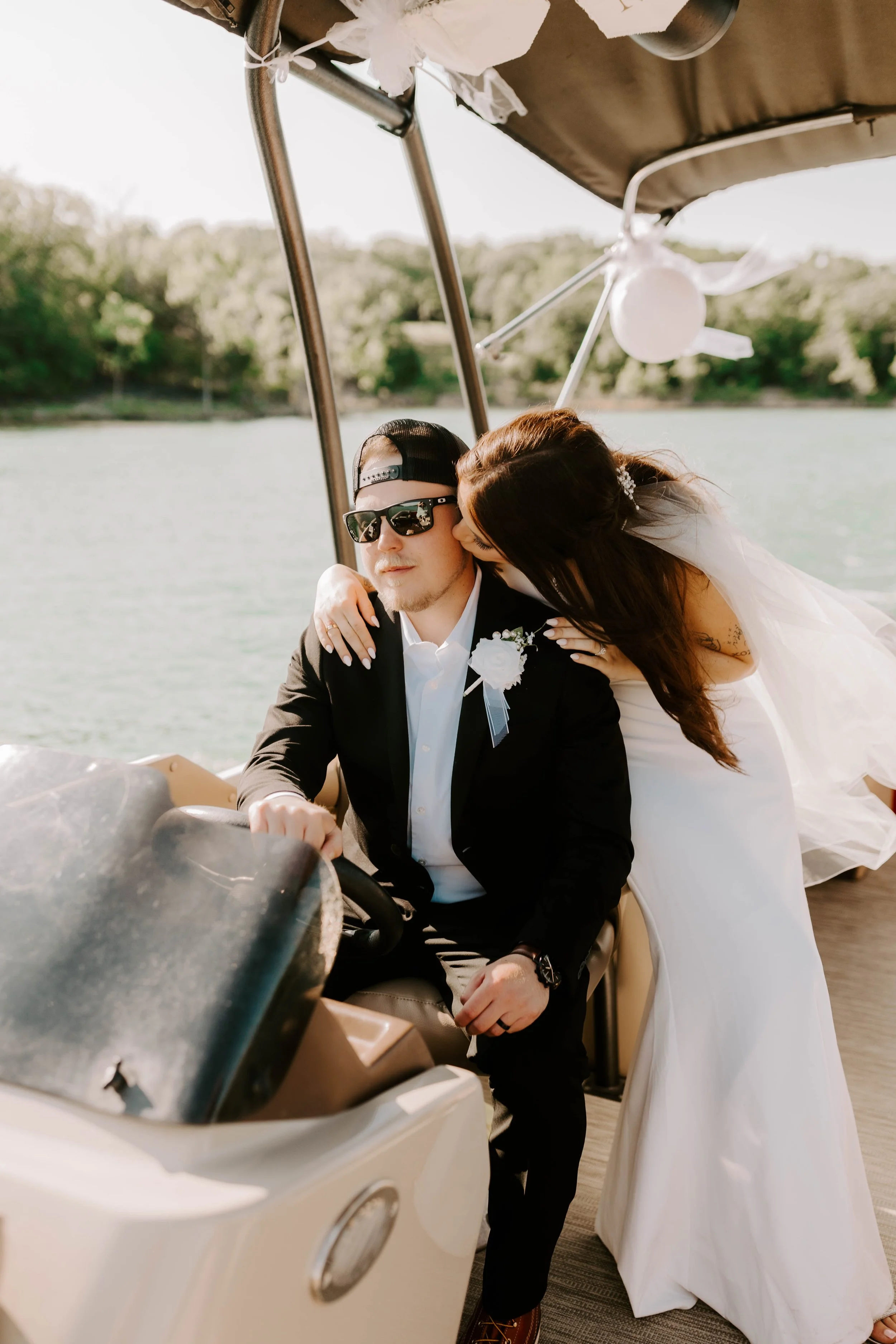 A man in a black suit and sunglasses sitting on a golf cart. A woman in a white dress is leaning over him, giving him a kiss on the cheek. The background shows a river and trees.