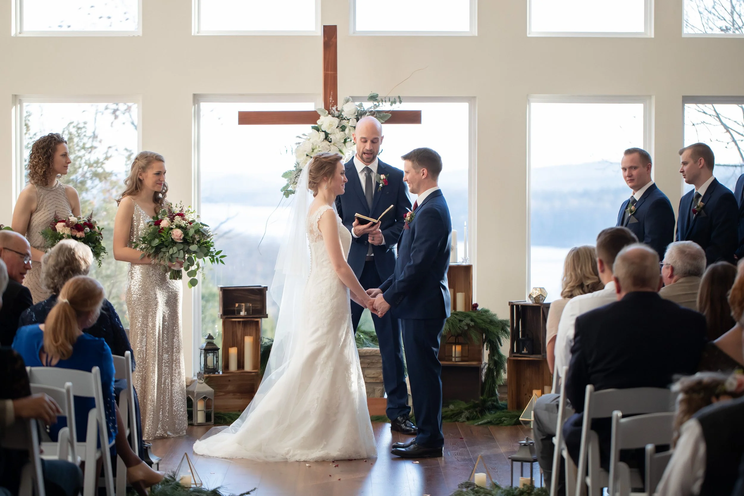A couple gets married during a ceremony in a bright, modern church with large windows and a wooden cross behind them. Bridesmaids and groomsmen stand nearby, and guests sit in white chairs watching the ceremony.