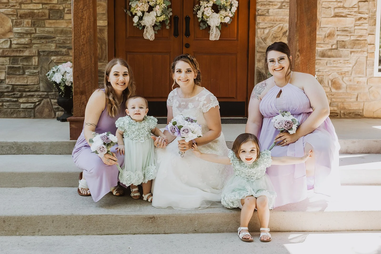 A bride and four women, possibly bridesmaids, sitting on steps in front of a stone and wood door at a wedding. The women are holding bouquets with roses, and two young girls are also present, one standing and one sitting, both dressed in light-colored dresses with floral details.