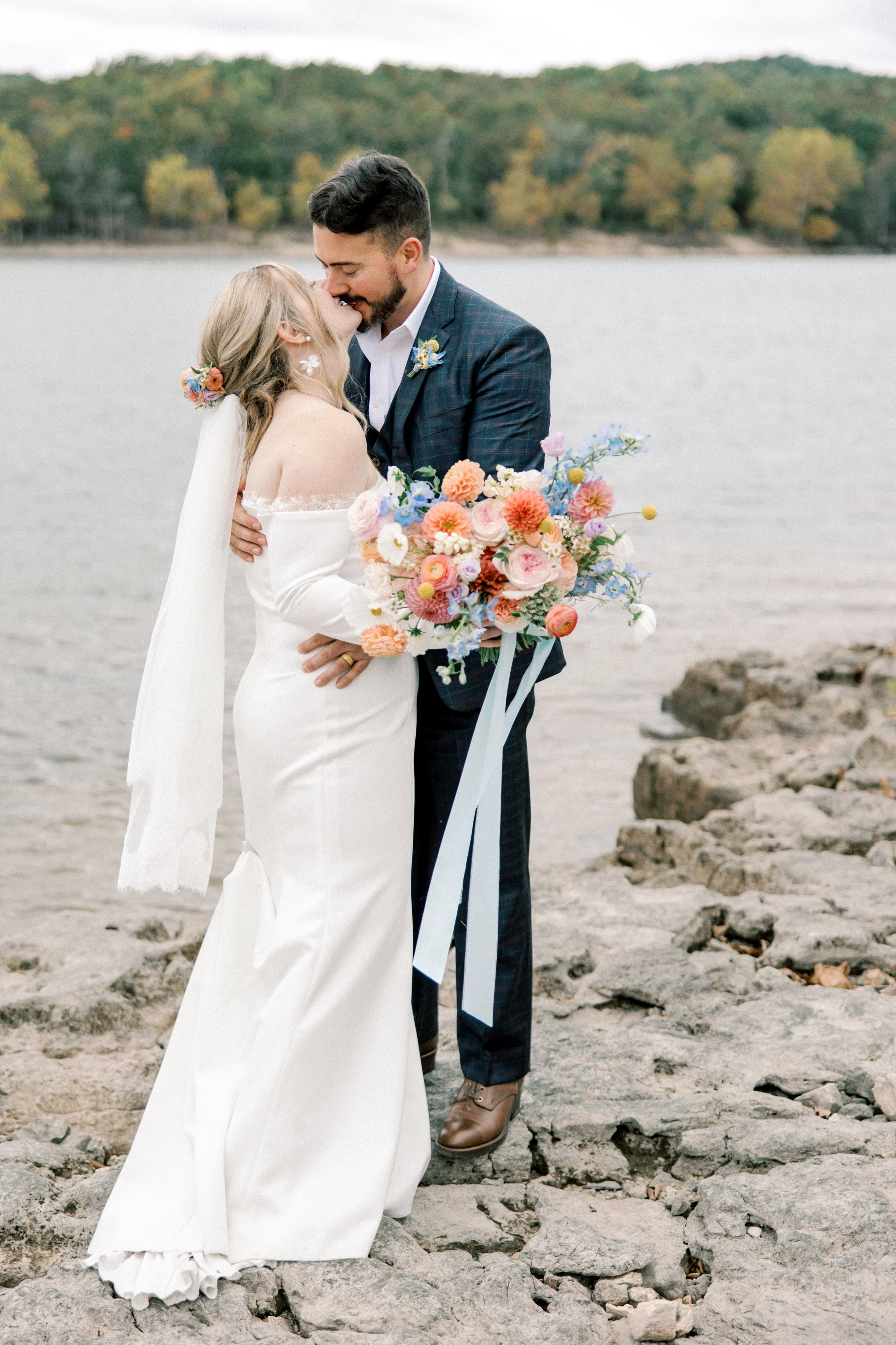 A bride and groom kissing on a rocky beach with water and trees in the background, the bride holding a colorful bouquet of flowers.