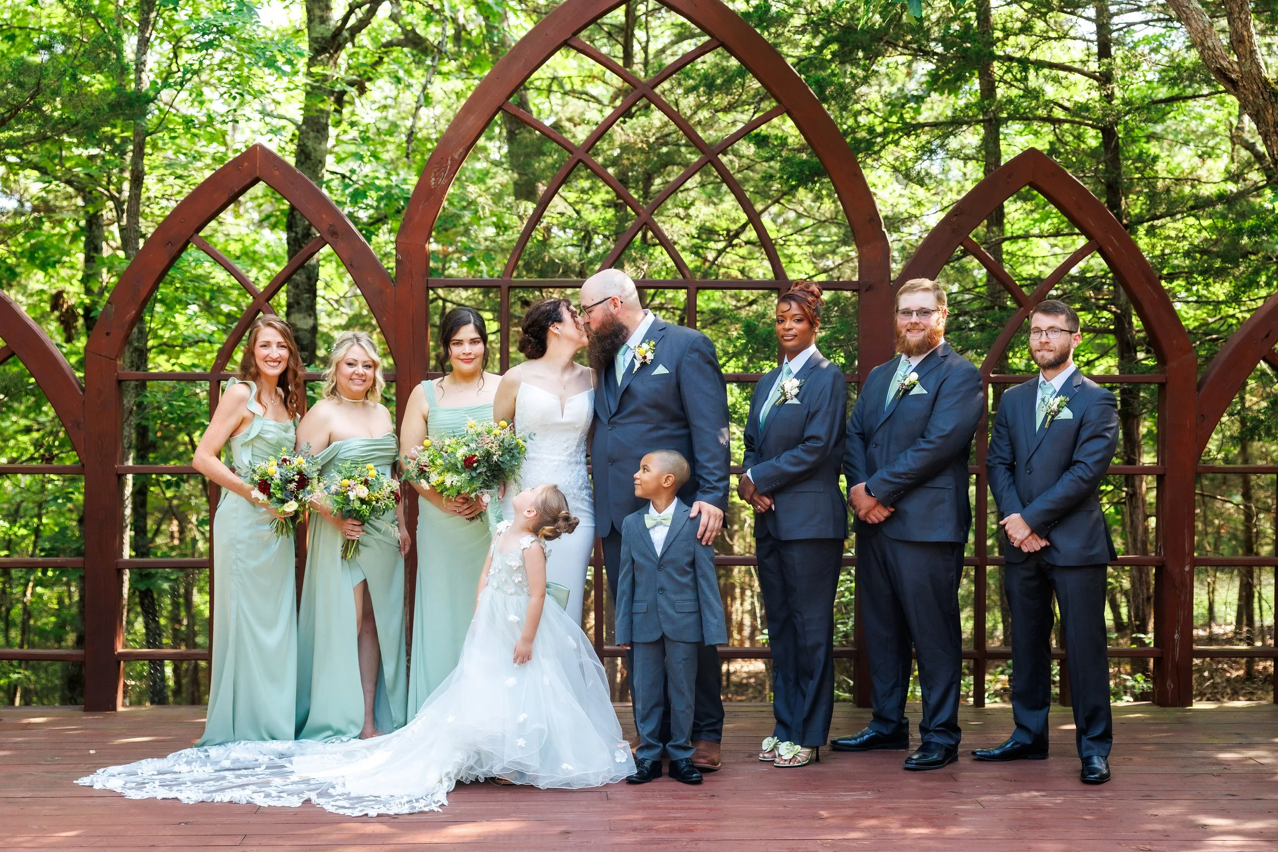 A group of wedding party members, including bride and groom, standing on a wooden platform in a forest, with some members holding bouquets and children in front, sharing a kiss.