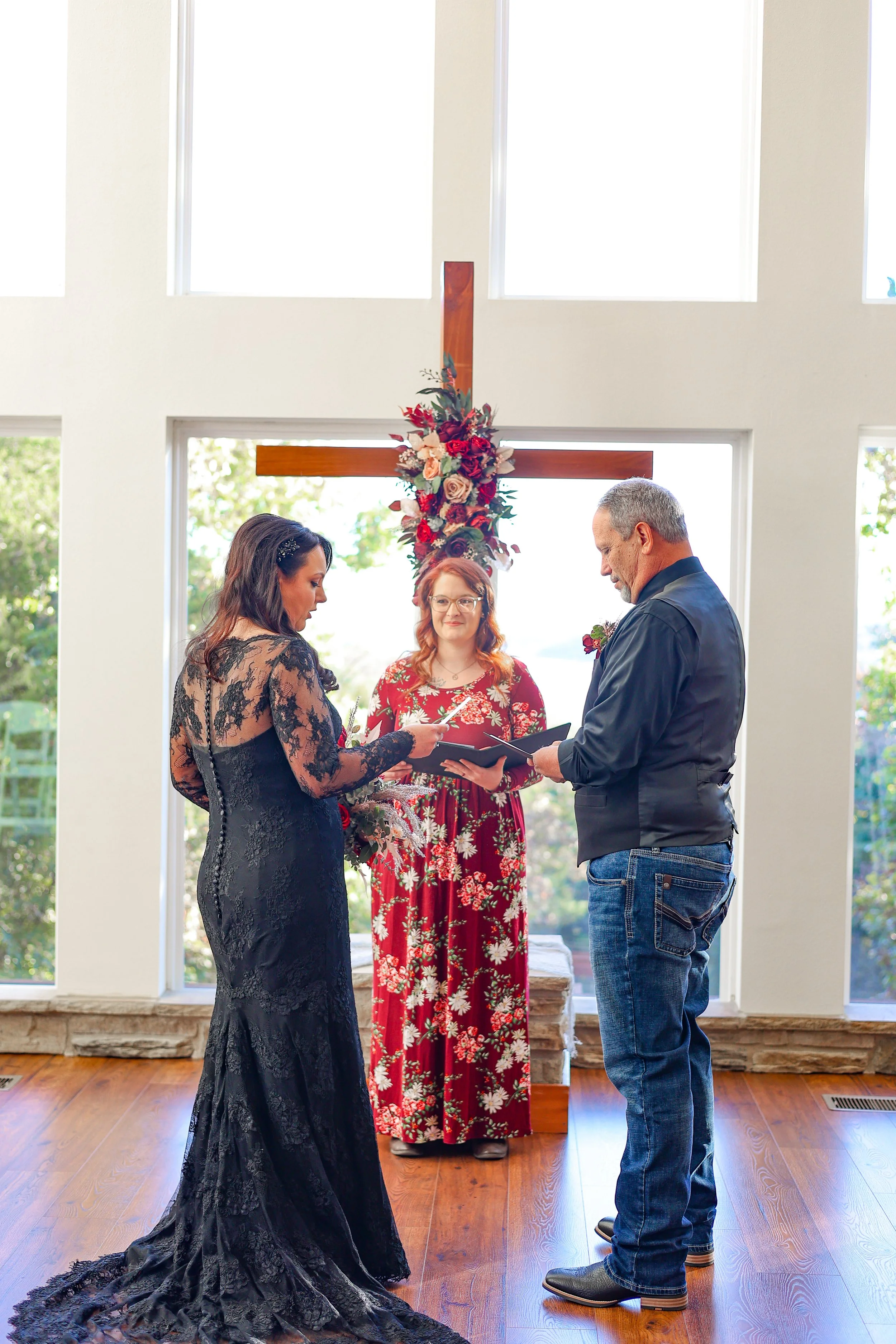 A couple is getting married in a church, with a woman in a black lace dress and a man in jeans and a dark shirt holding hands in front of a priest and an officiant. There is a large wooden cross decorated with flowers in the background, and sunlight 