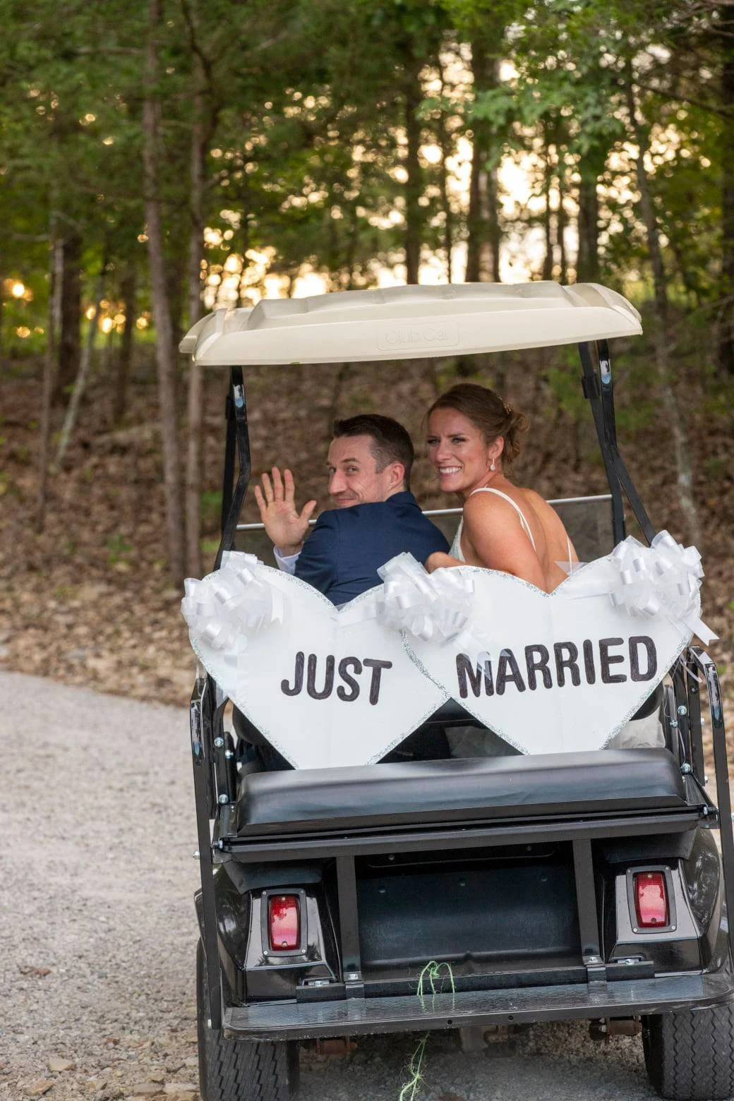 A newlywed couple on a golf cart with a “Just Married” sign, smiling and waving, surrounded by trees at sunset during their wedding celebration.